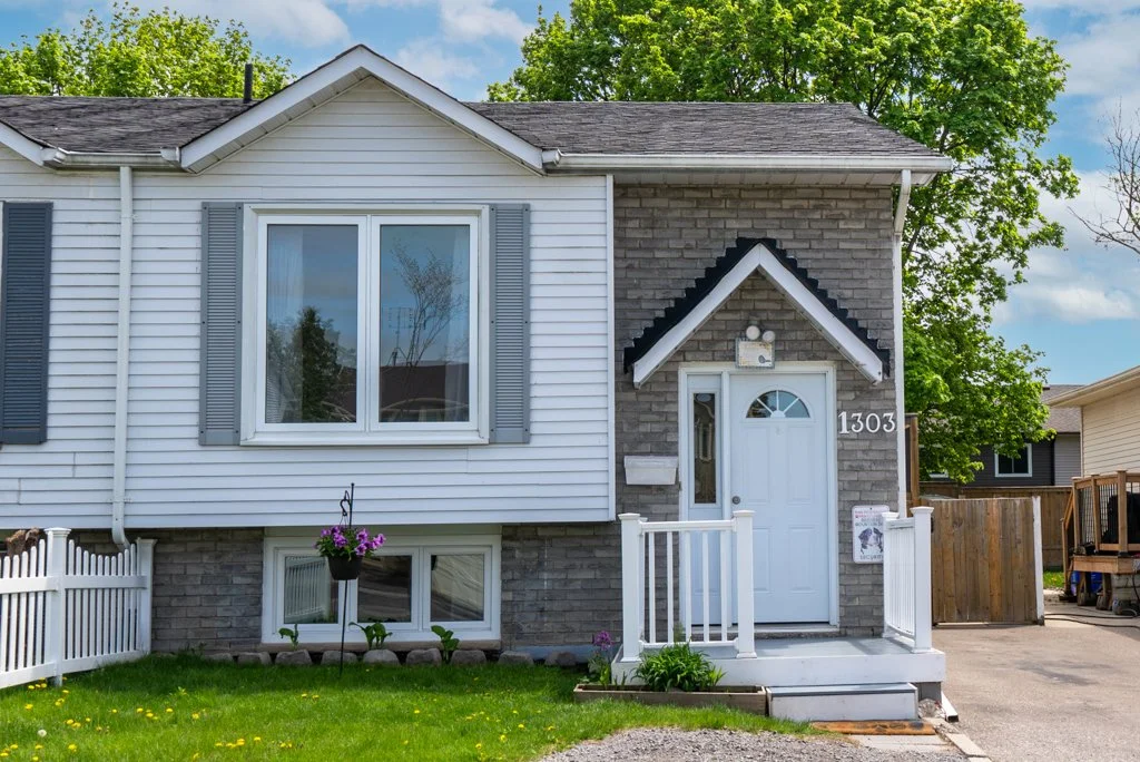 Front view of a two-story house with white siding and gray brick, featuring a white front door with sidelights, a small porch with white railings, large double-hung window with shutters, and a manicured lawn with flowers and hanging basket.