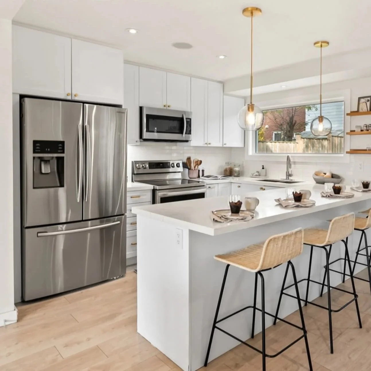 Modern kitchen with white cabinets, stainless steel refrigerator, microwave, and stove. An island with seating for three, set with breakfast items, and pendant lights overhead. Large window above the sink with a view of a backyard.