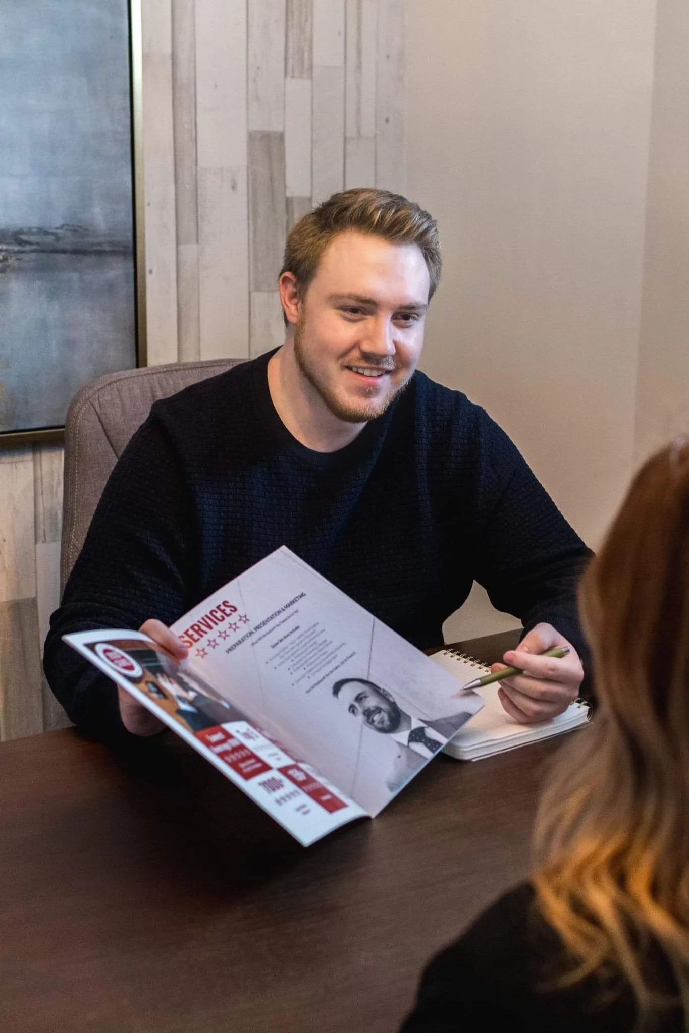 A young man with light brown hair, beard, and wearing a black sweater, sitting at a table during an interview or consultation, holding a brochure or magazine with the words 'SERVICES' visible.