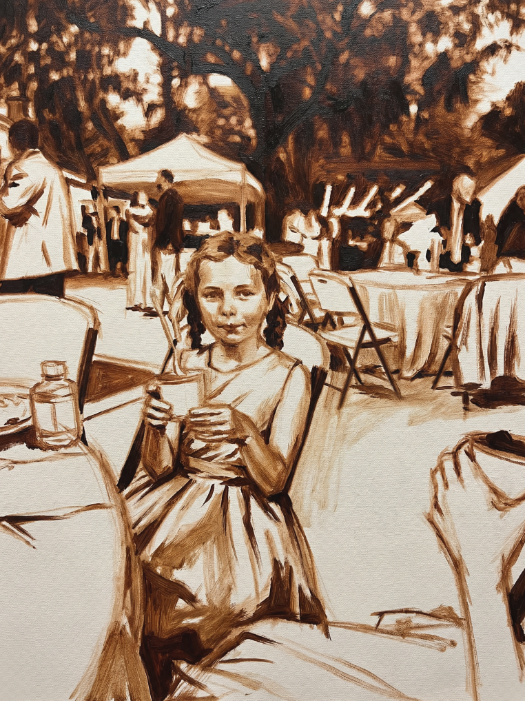 A girl sitting at a table outdoors during a gathering or event, holding a cup and looking at the camera, with tables and people in the background under a tree.