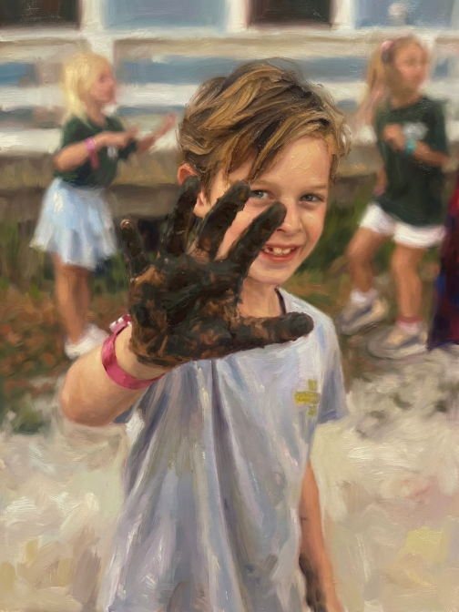 Young girl with short hair smiling and holding up a hand covered in dark paint or mud in an outdoor setting with other children in the background.