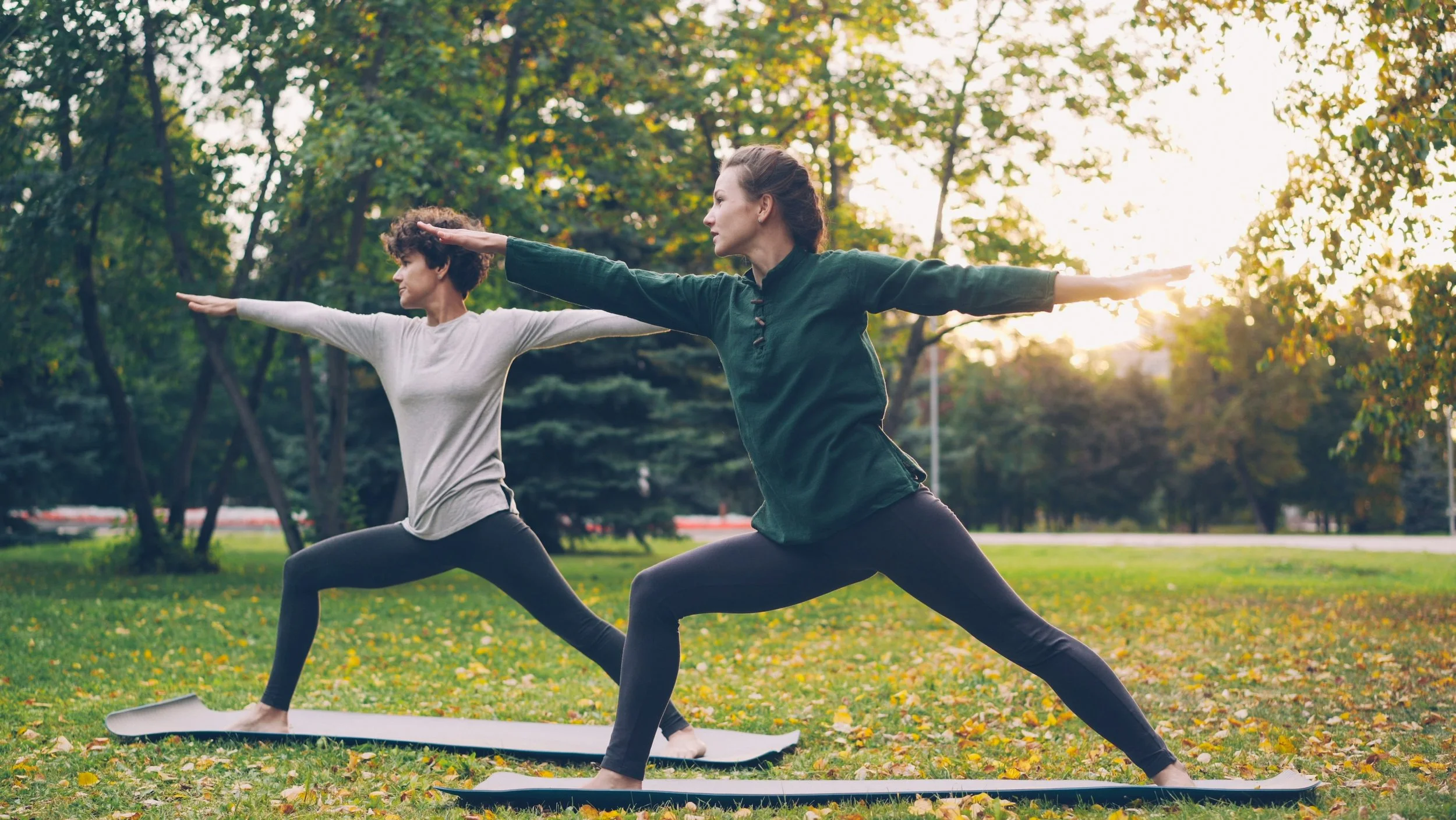 Two women practicing balancing yoga poses together in an autumn park, focusing on wellbeing and calm.