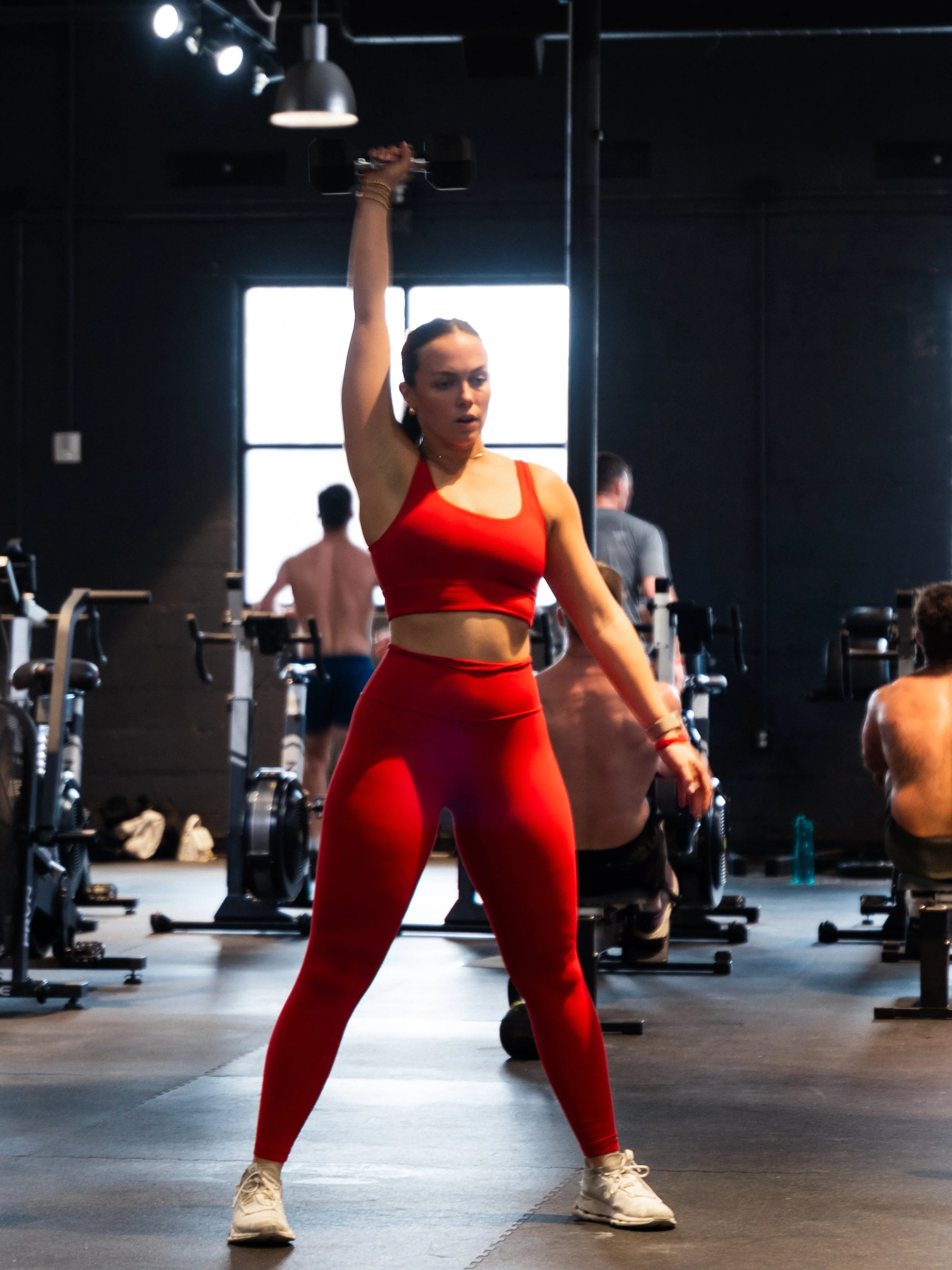 A woman in red workout clothes is lifting a dumbbell overhead in a gym. There are other people working out in the background.