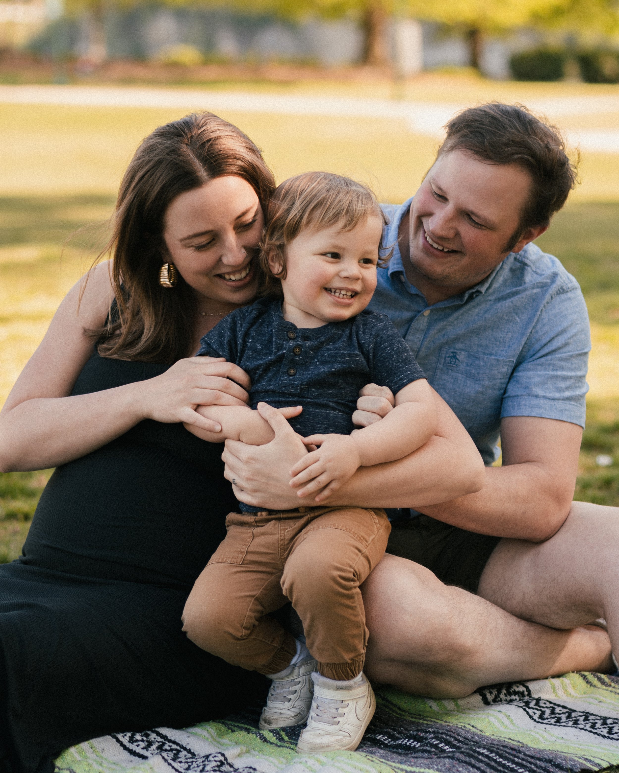 Happy family of three sitting on a blanket outdoors in a park, with trees and grass in the background; the mother and father are holding their young child, smiling and laughing together.