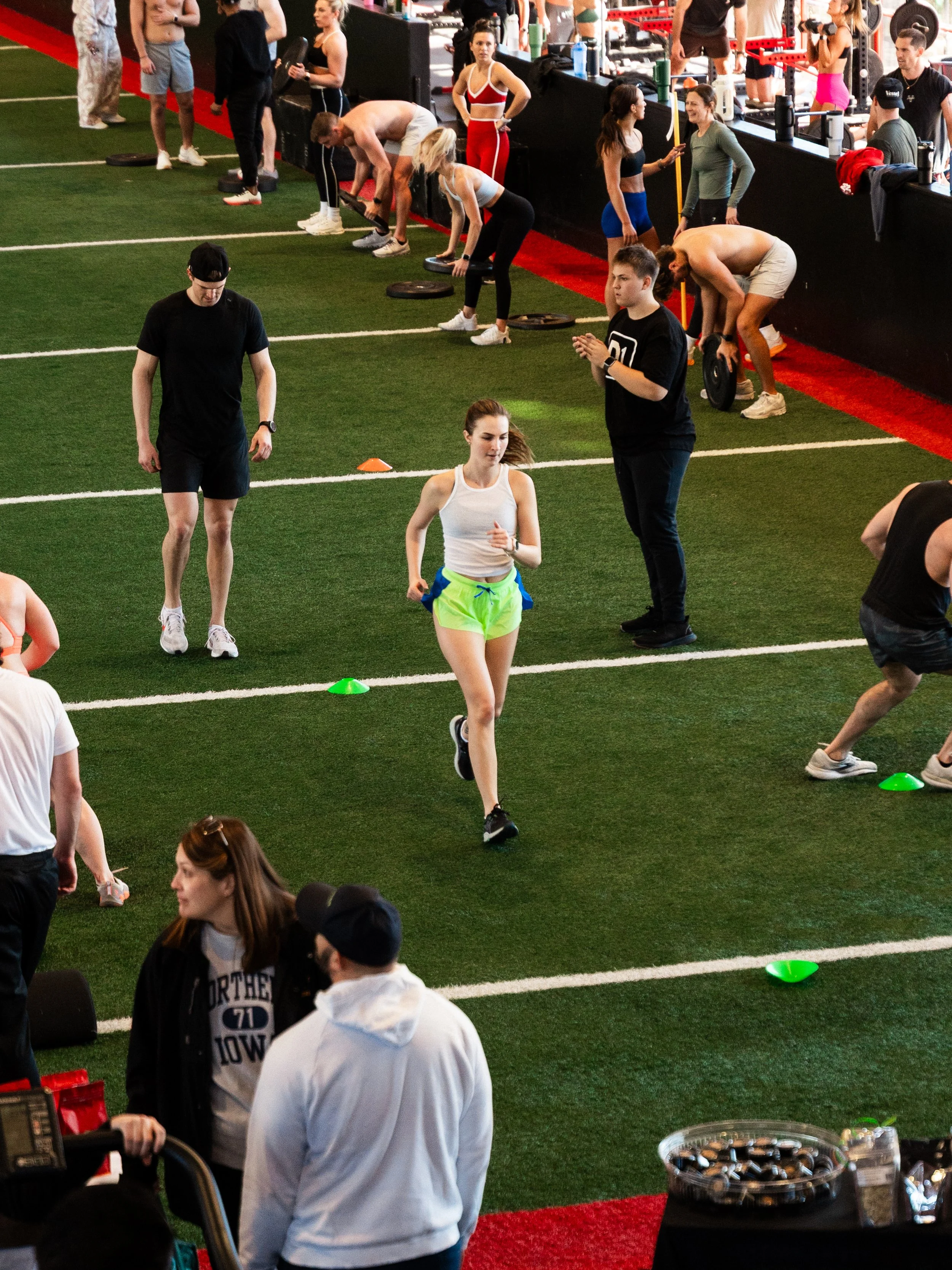 People exercising on a turf training area with weights and cones, some lifting weights, others running, and some standing around watching or giving instructions, in an indoor gym or fitness event.