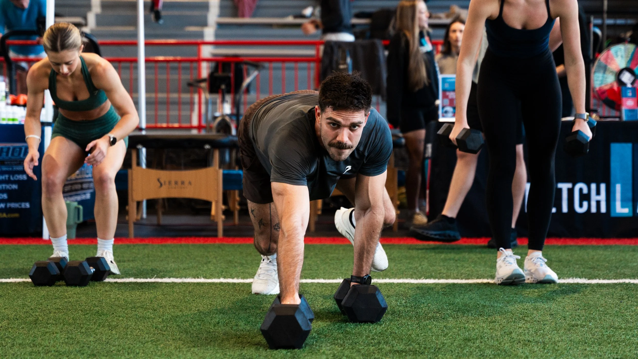 A man performing a push-up with dumbbells on a gym floor, surrounded by women with dumbbells, in an indoor sports facility.