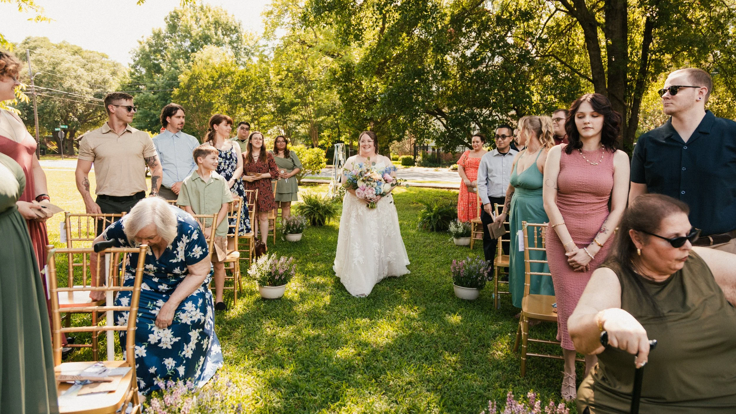 A bride walking down the aisle at an outdoor wedding ceremony surrounded by guests sitting on gold chairs, lush green trees, and potted flowers on a sunny day.
