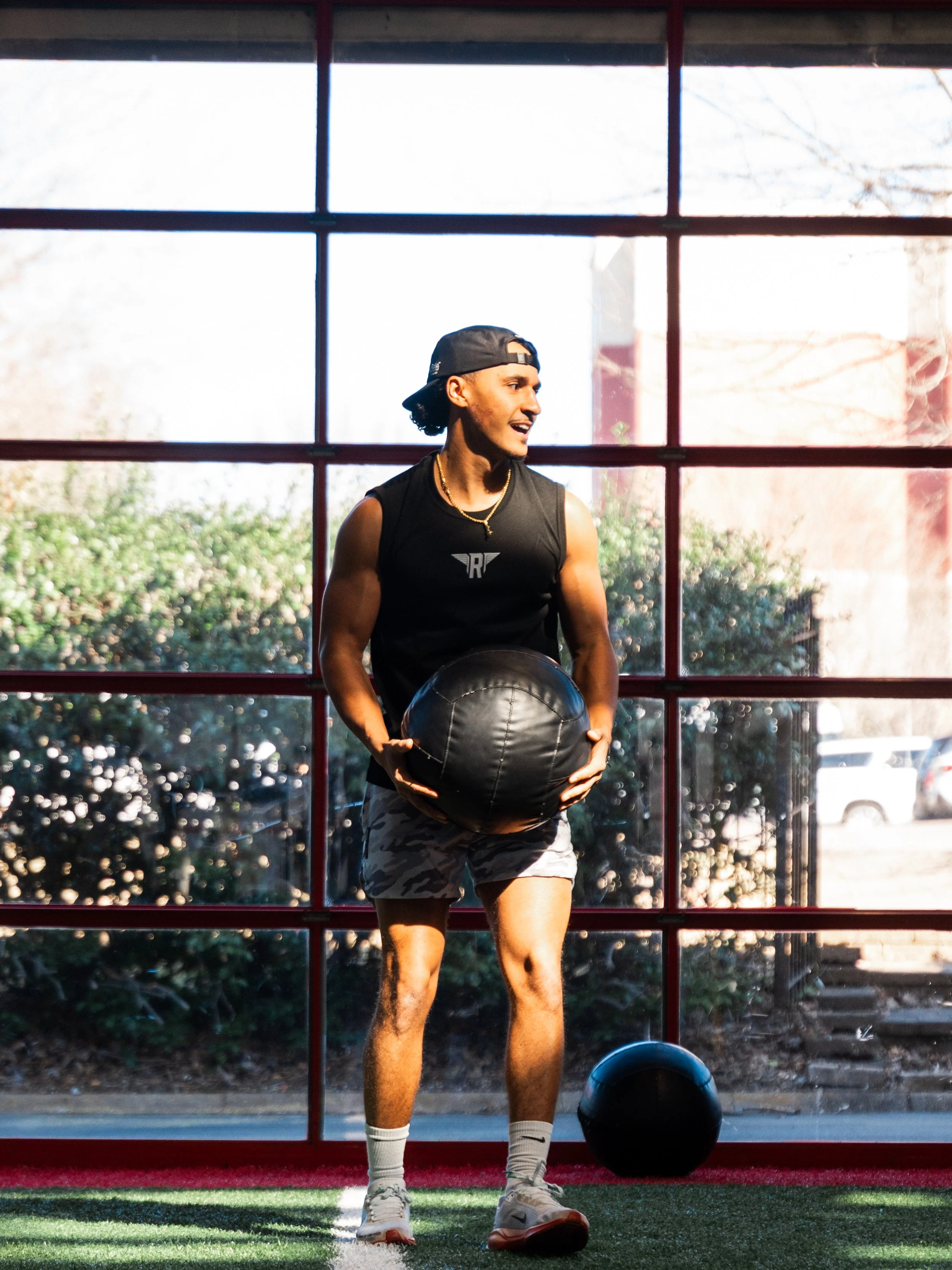 A young man in athletic clothing and a backwards cap holding a medicine ball in a gym with large windows, sunlight outside, and a black helmet on the floor.