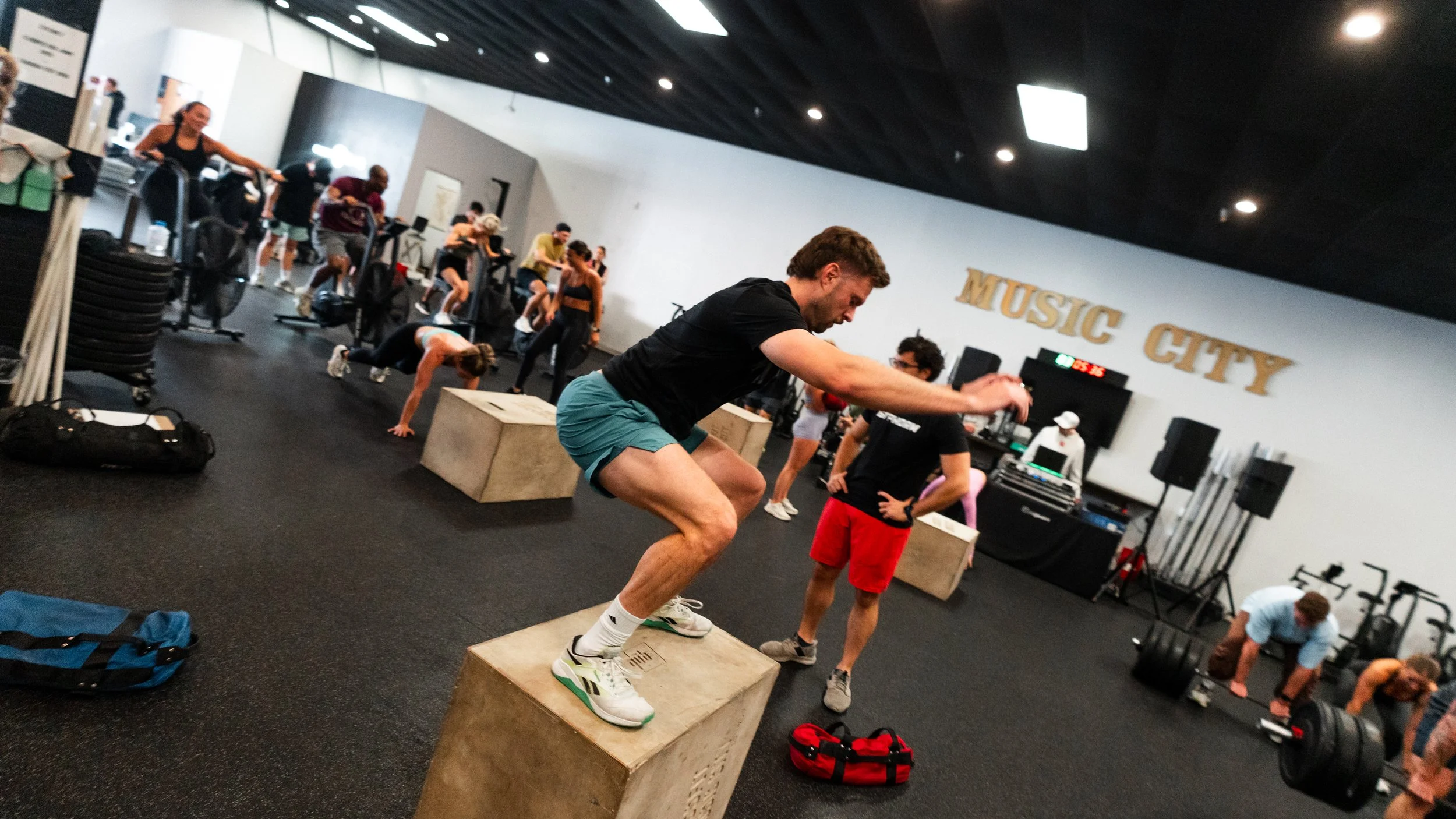 People exercising in a gym, with a man jumping onto a plyometric box, surrounded by others doing various workouts.
