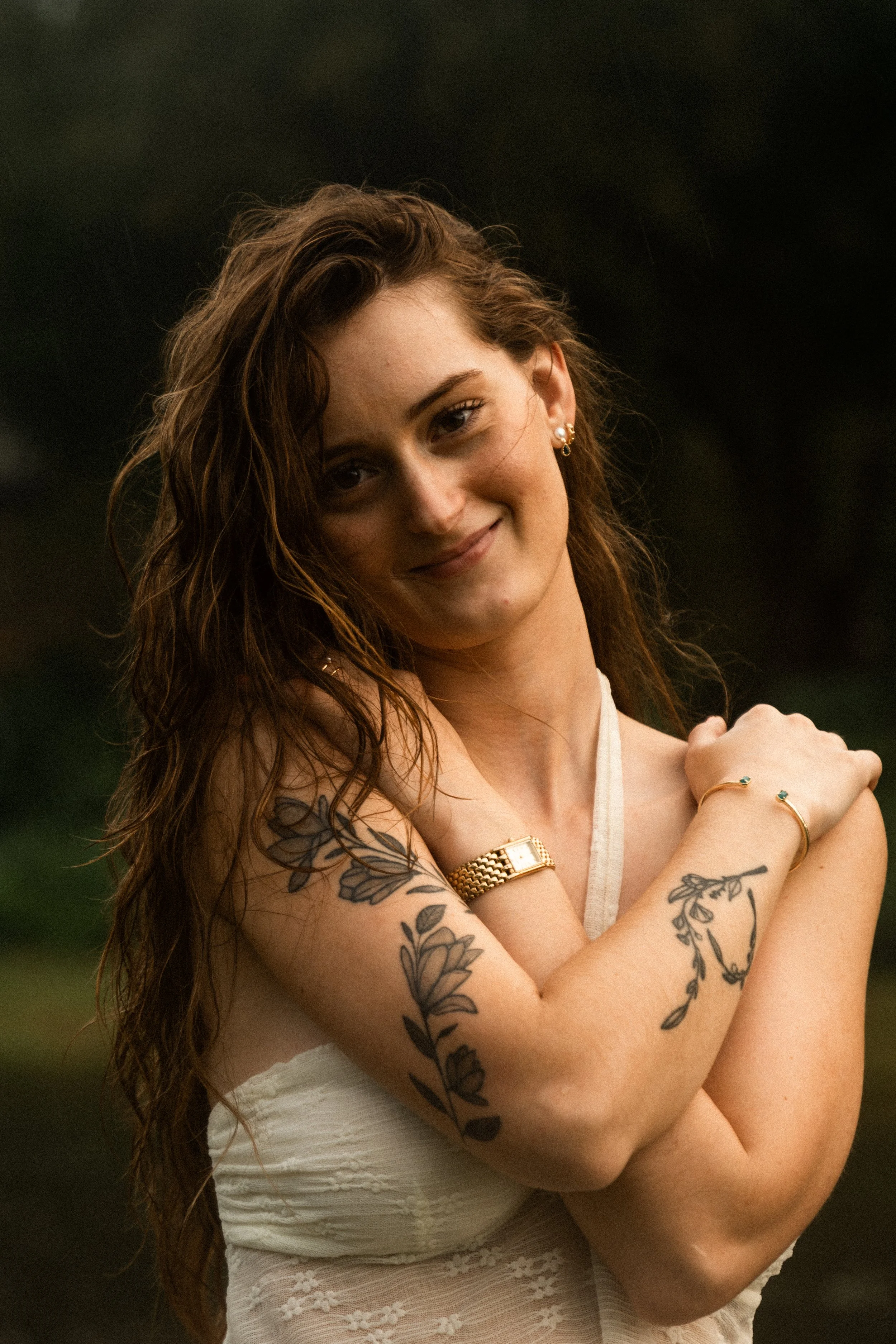 A young woman with long wavy brown hair and multiple tattoos on her arms is smiling and looking at the camera. She is wearing earrings, a gold watch, and bracelets. She is dressed in a sleeveless white lace top and is outside with a dark, blurred background.