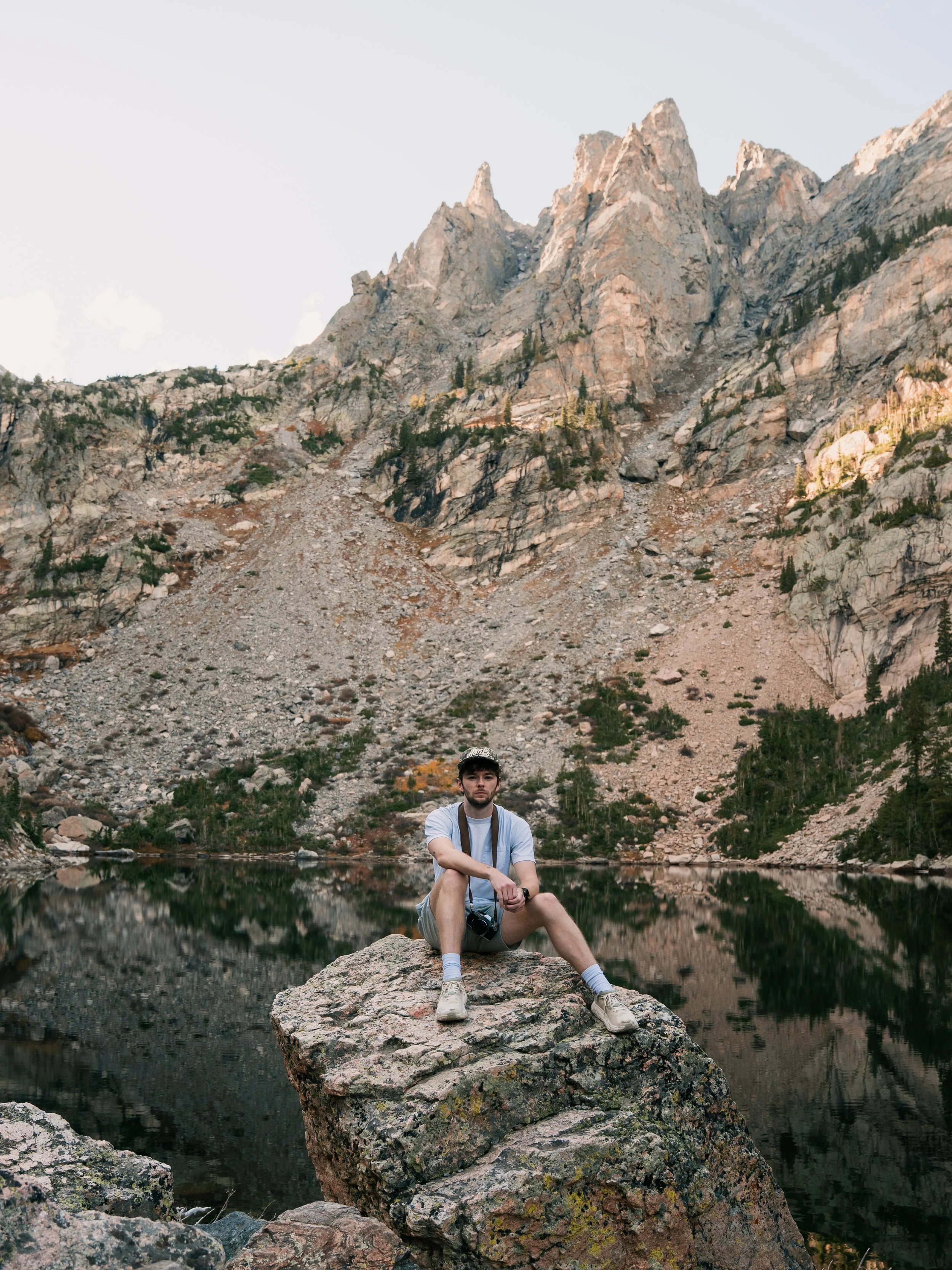 A man sitting on a large rock near a body of water with mountain peaks in the background.