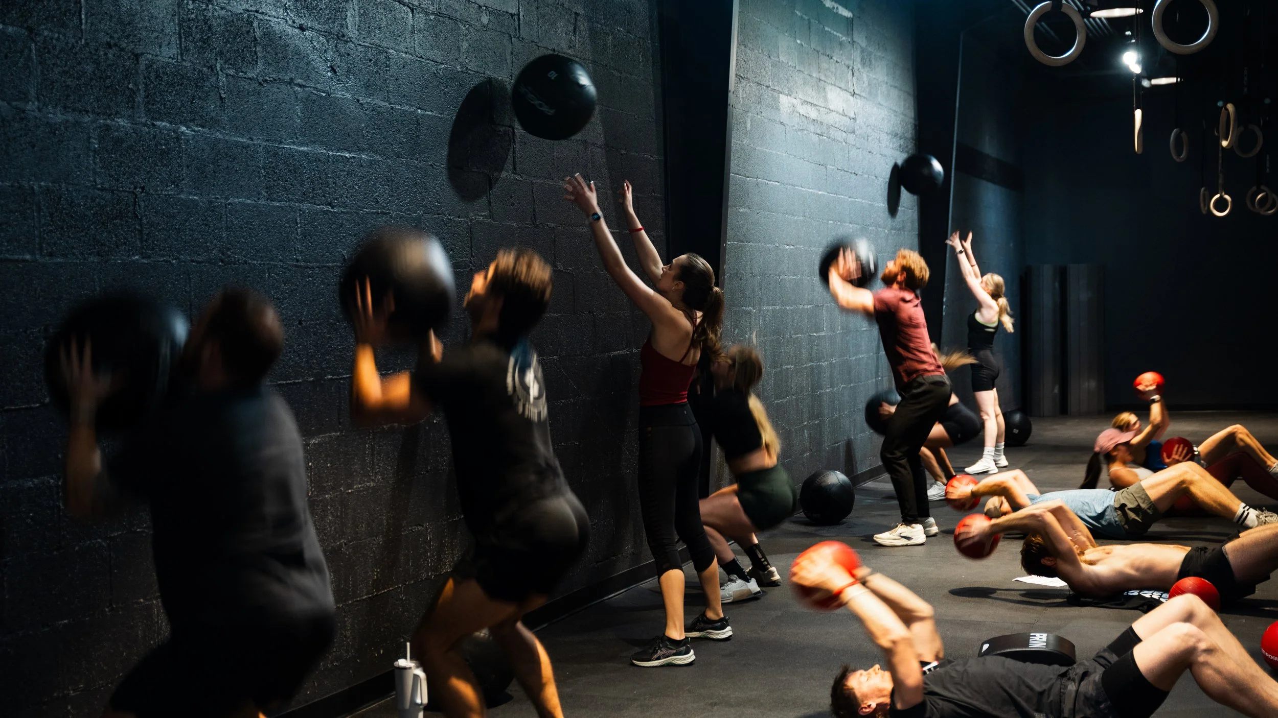 People participating in a fitness class doing wall ball exercises, using medicine balls against a black wall in a gym.