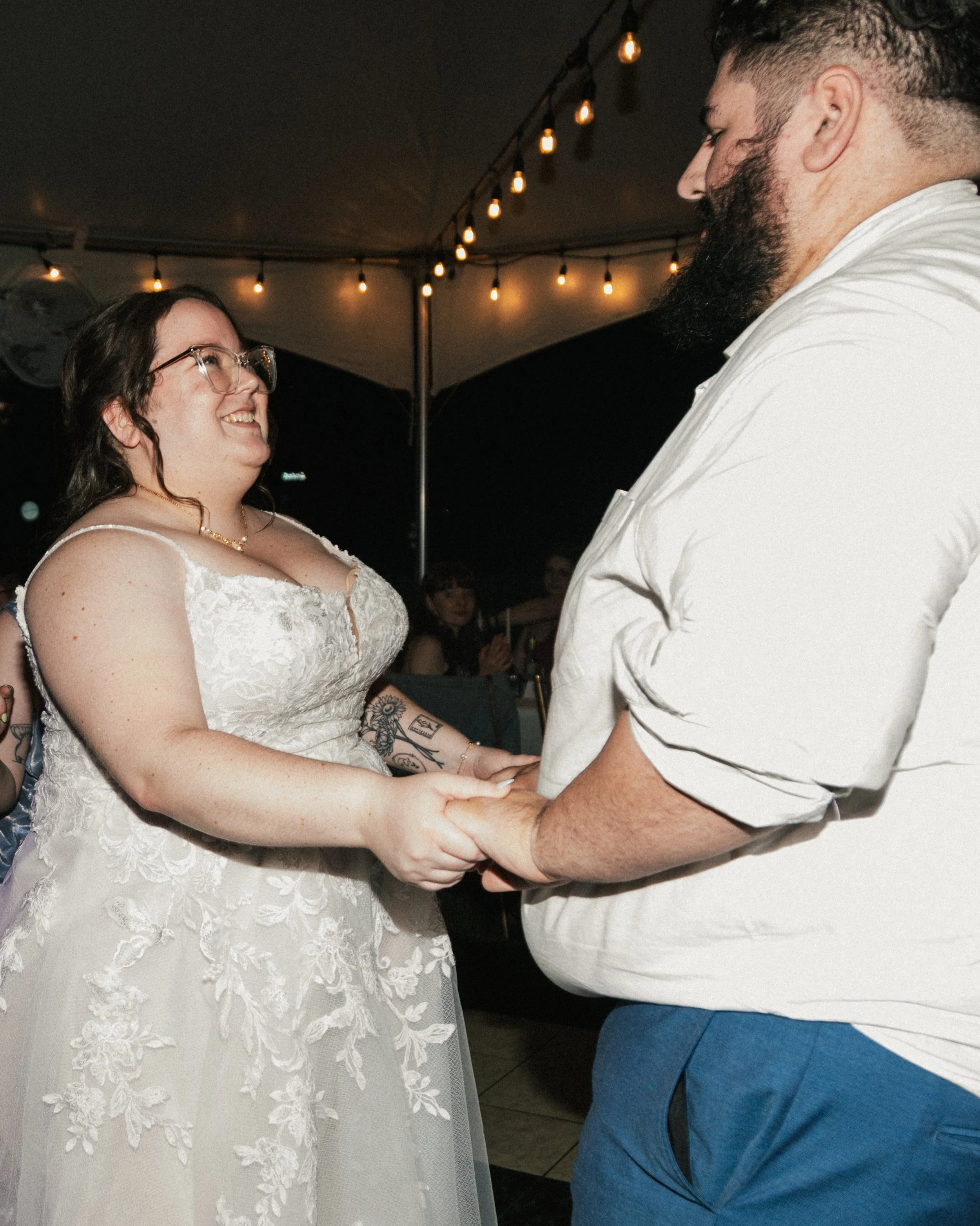 A couple is holding hands during a wedding dance, with the bride wearing a white lace wedding dress and glasses, and the groom wearing a white shirt and blue pants. String lights decorate the ceiling in the background.