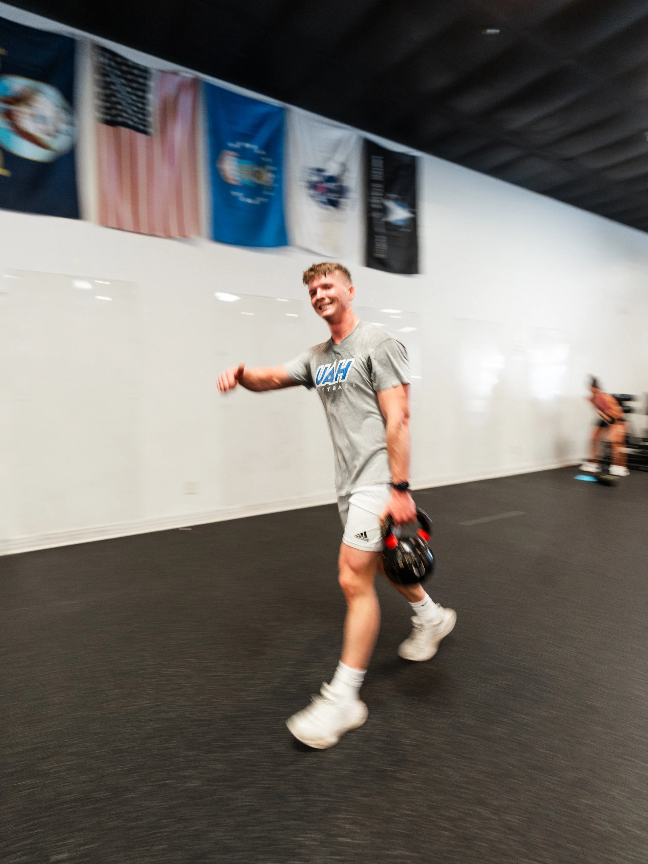 A young man in athletic clothing walking in an indoor gym with flags hanging on the wall behind him, holding a kettlebell in his right hand, smiling.