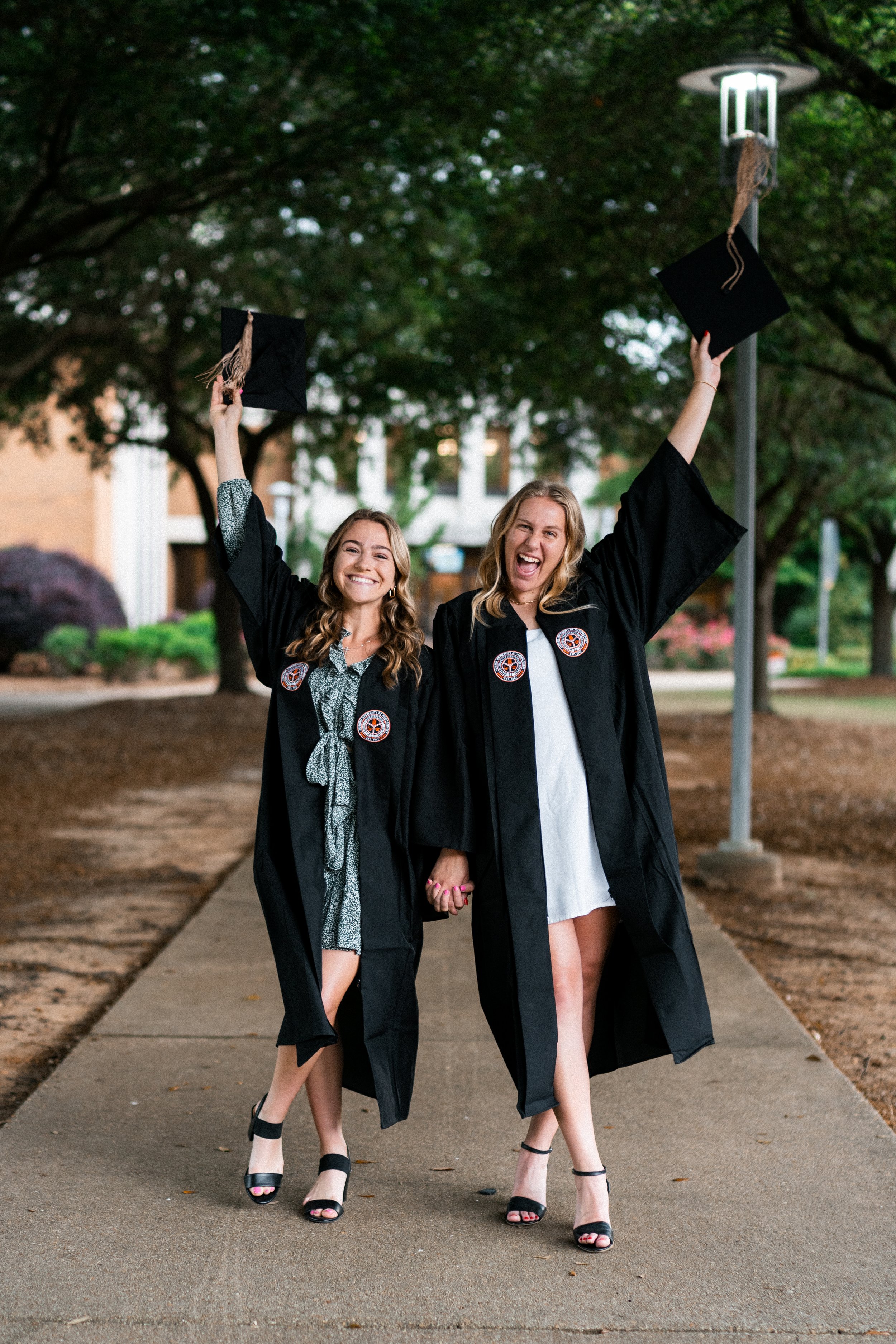 Two women in black graduation gowns celebrating outdoors, holding hands, with graduation caps raised in the air, smiling and walking on a sidewalk under trees at dusk.