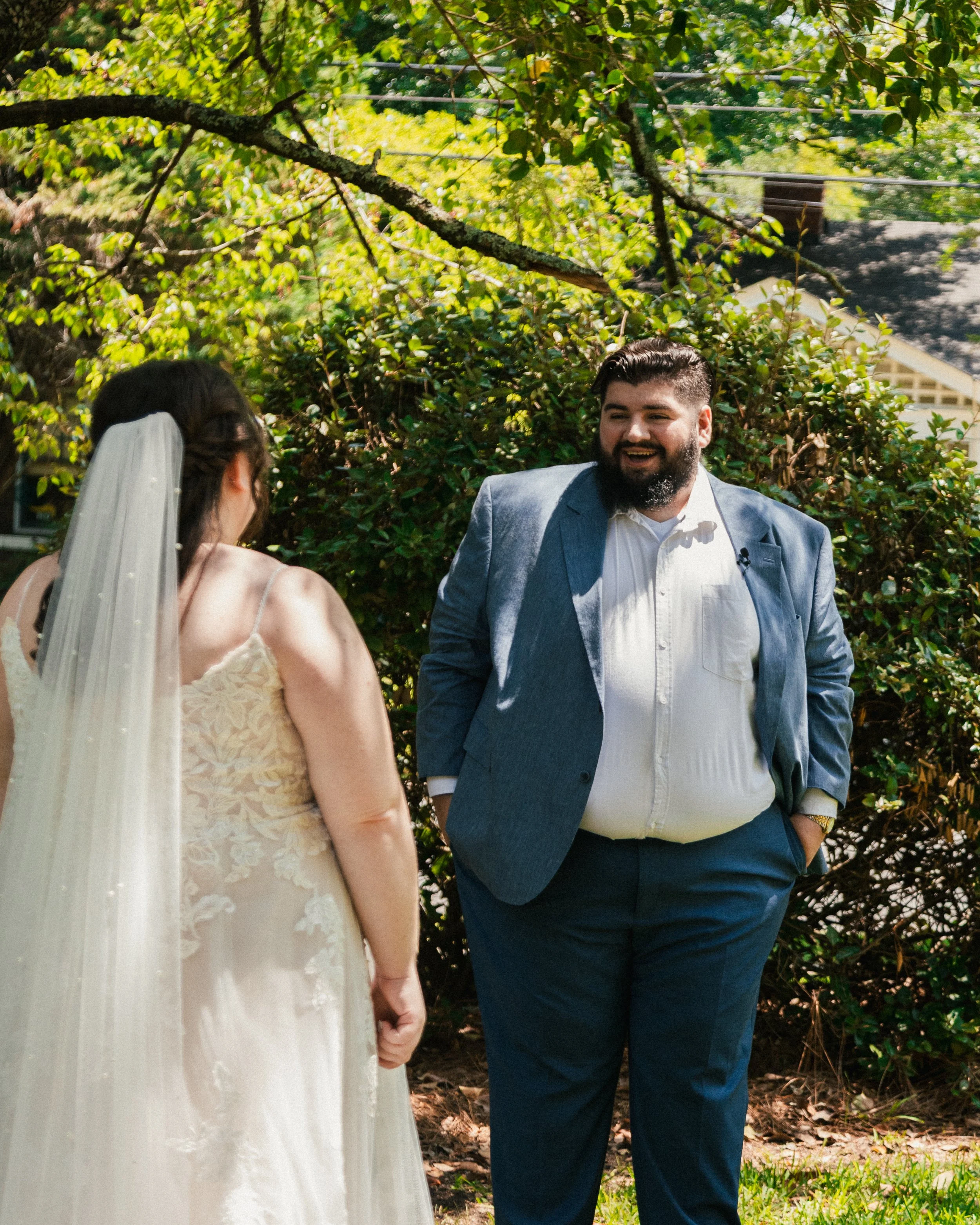 A bride wearing a white dress and veil holding hands with a groom in a blue suit, smiling outdoors surrounded by green trees and bushes.