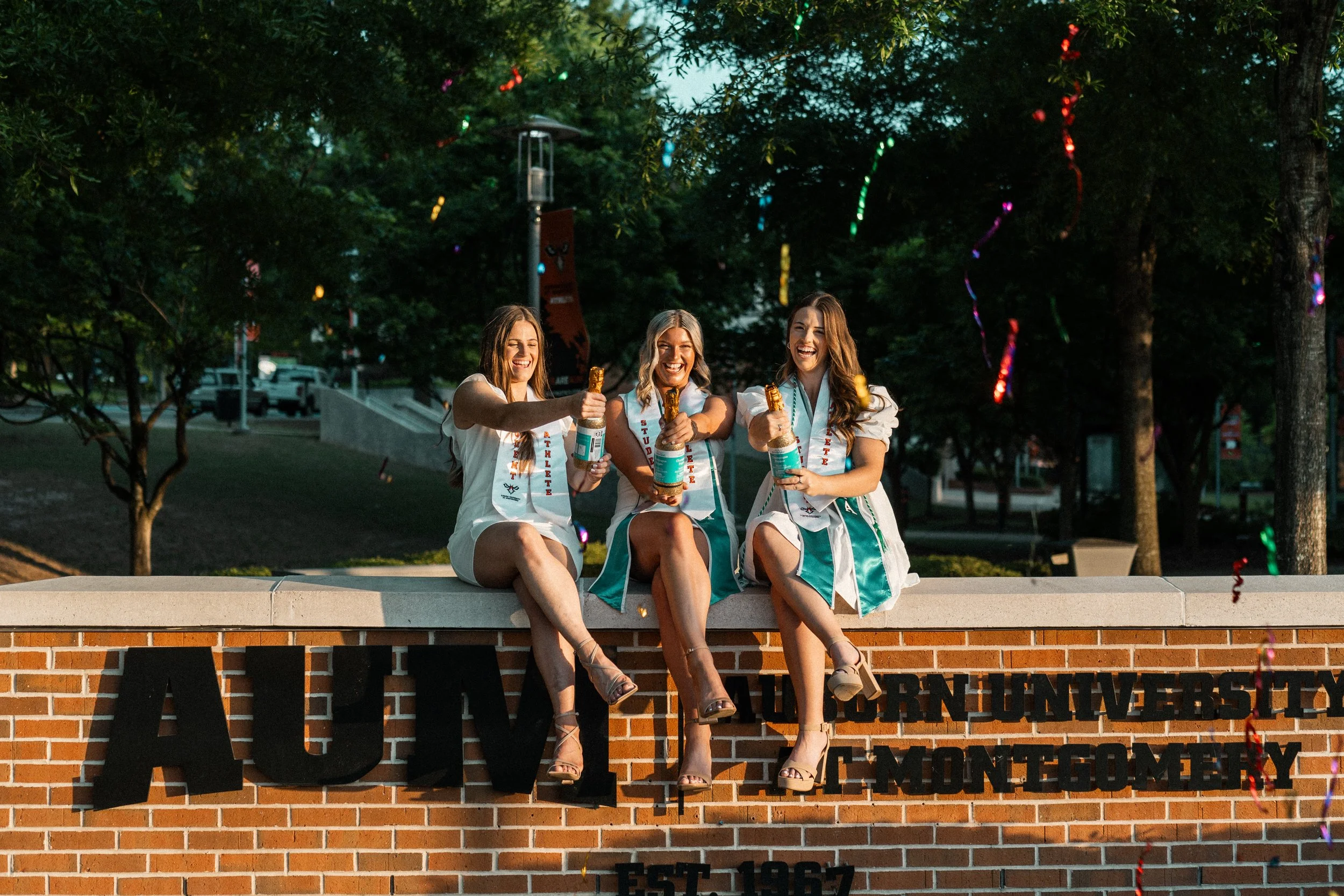 Three young women, dressed in white and teal, sitting on a brick wall at an outdoor event, holding bottles of champagne, smiling, with trees and colorful string lights in the background.