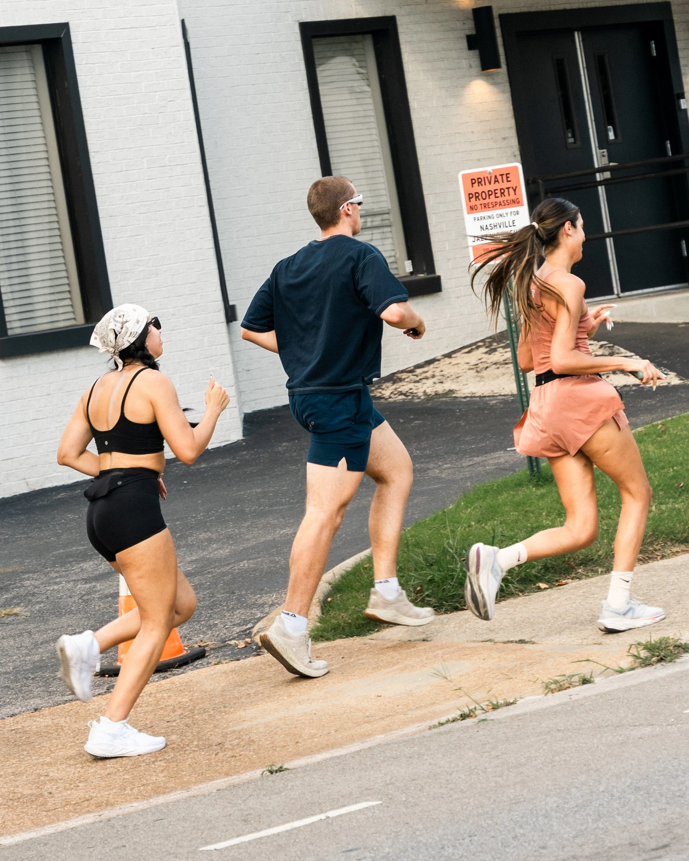 Three people running along a sidewalk in front of a brick building with a "Private Property" sign. Two women and one man, all dressed in athletic clothing and sneakers, are mid-stride.