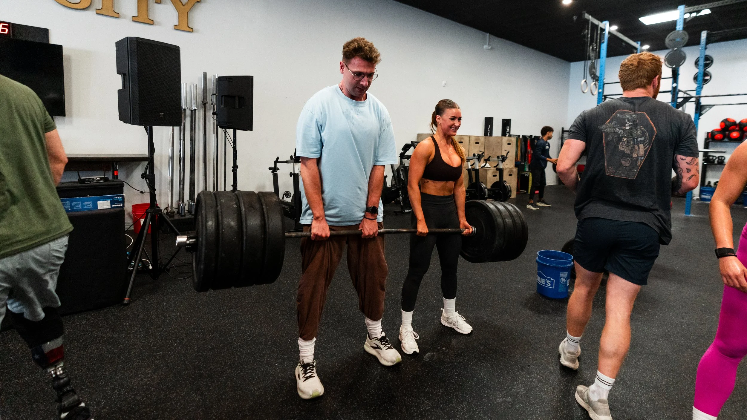 People lifting weights in a gym, with one man in a light blue t-shirt and brown pants holding a heavy barbell, appearing focused, and a woman in a black sports bra and leggings smiling nearby. Other gym members are visible in the background.