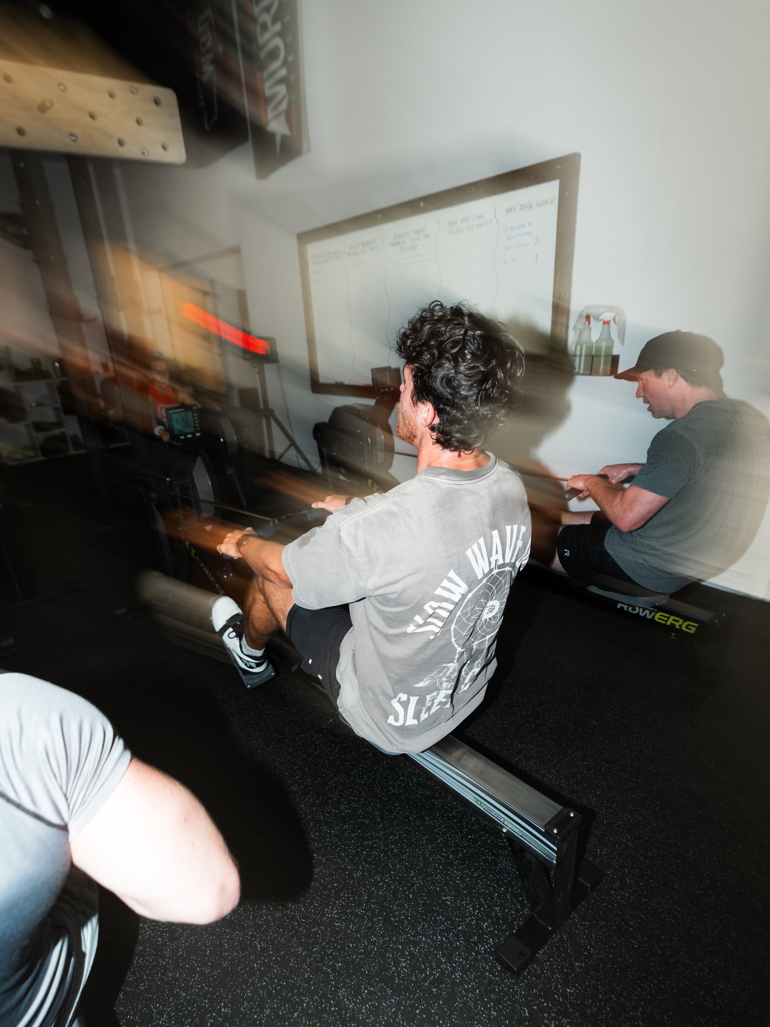 People working out on rowing machines in a gym, with a whiteboard on the wall behind them and a person wearing a gray T-shirt with text on the back.
