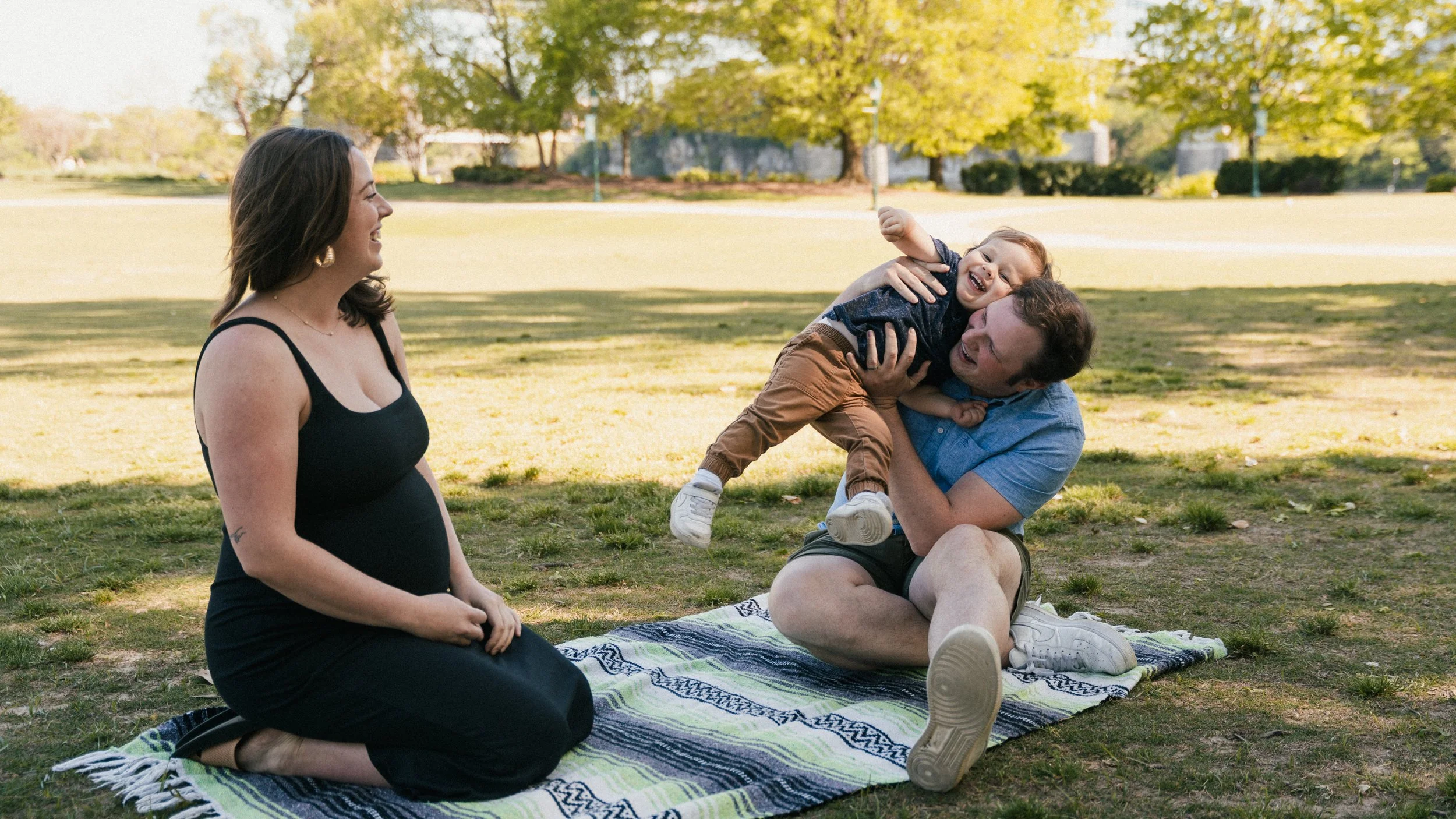A family enjoying an outdoor day in a park. A woman is sitting on a blanket with two children, one of whom is being playfully lifted in the air by a man. The scene is set under trees with green leaves.