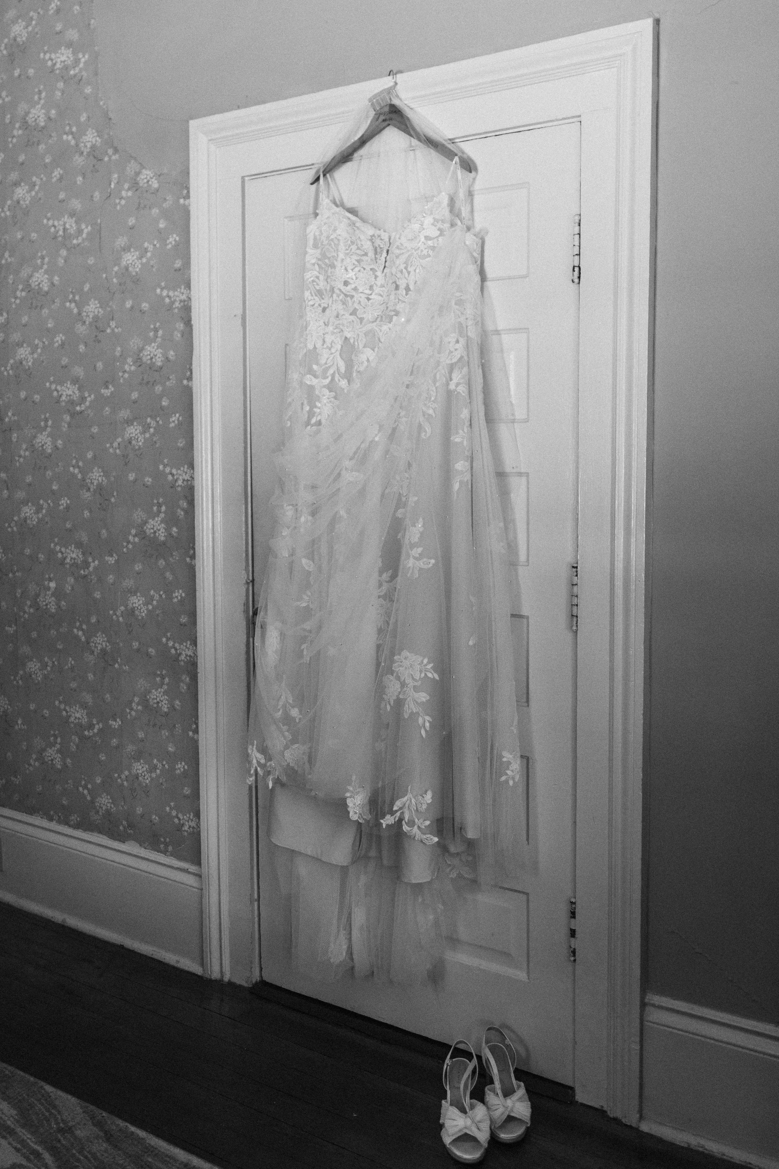 Wedding dress hanging on a hook inside a room with floral wallpaper on the left wall. White shoes with bows placed on the dark wood floor beneath the dress.