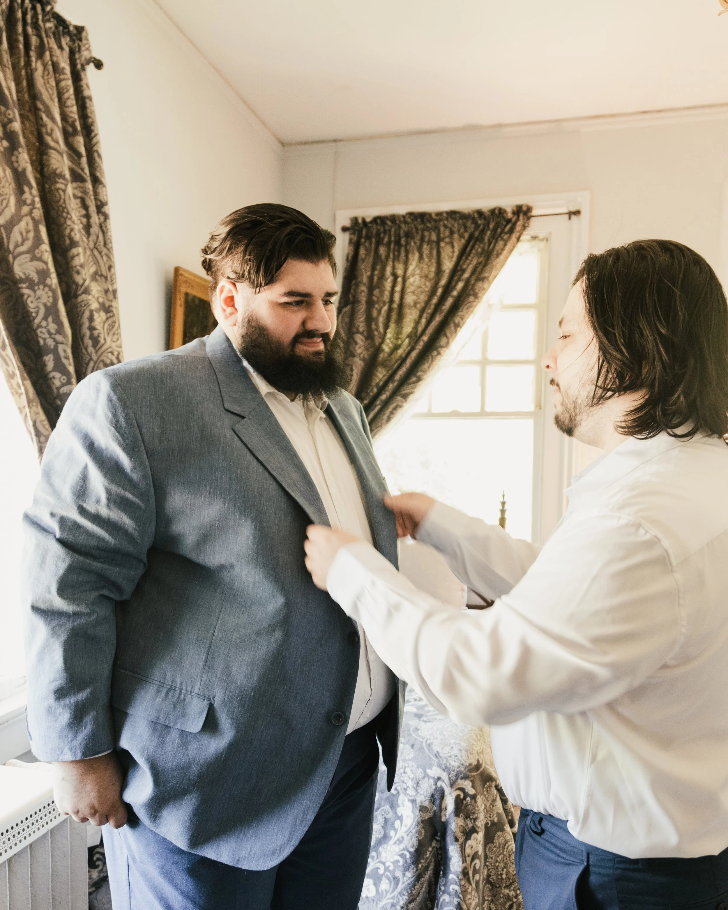 Two men in formal attire, one in a gray suit and the other in a white shirt and dark pants, are in a room with patterned curtains and a window. The man in the white shirt is adjusting the suit of the man in the gray suit.