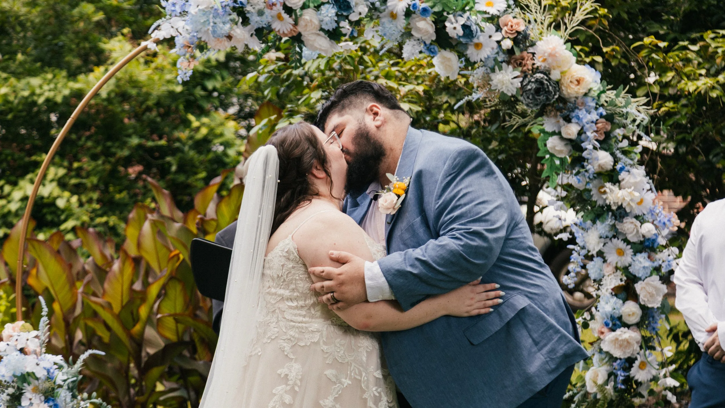 A newlywed couple sharing a kiss during their wedding ceremony outdoors, surrounded by a floral arch and greenery.