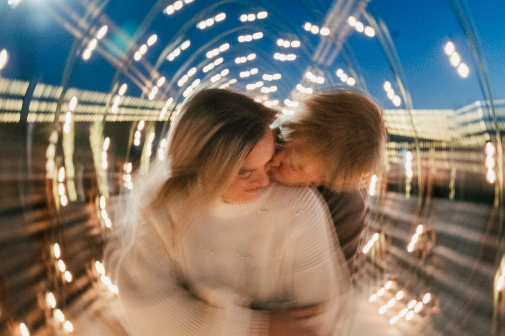 A young couple is embracing each other in a tunnel decorated with hanging string lights, with a blue evening sky visible above.