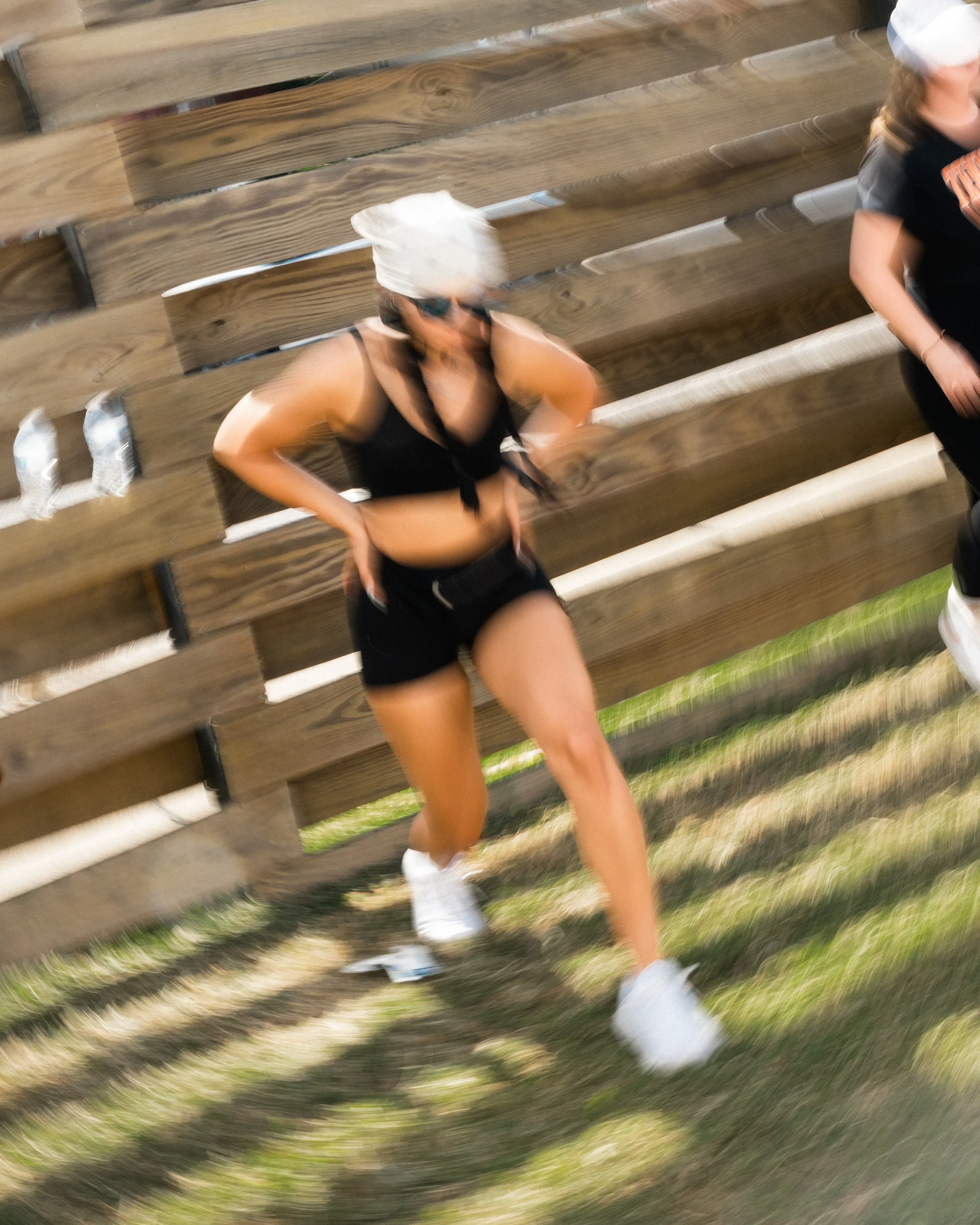 A woman in black athletic clothing, white cap, and sunglasses walking past a wooden fence with three water bottles on a bench, with grass and pathways visible.