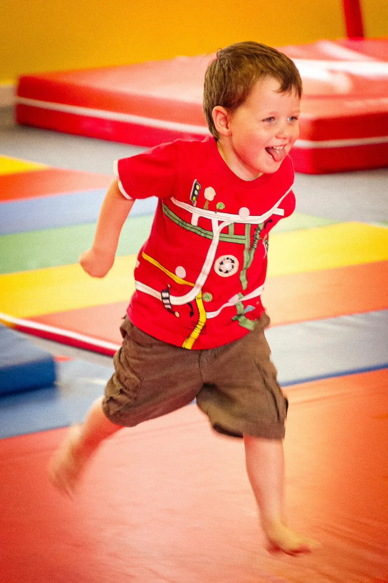 A young boy in a red shirt and brown shorts running and smiling on a colorful gym mat.
