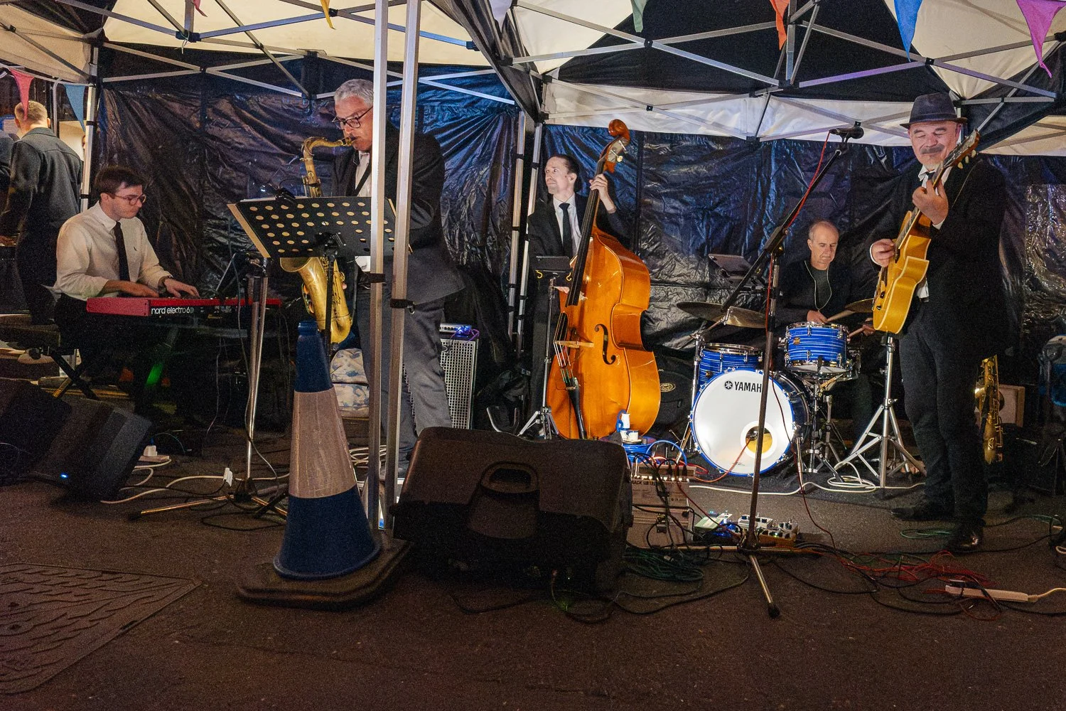 Five-piece jazz band playing keyboard, saxophone, double bass, drums and guitar under a gazebo on George Street, St Albans