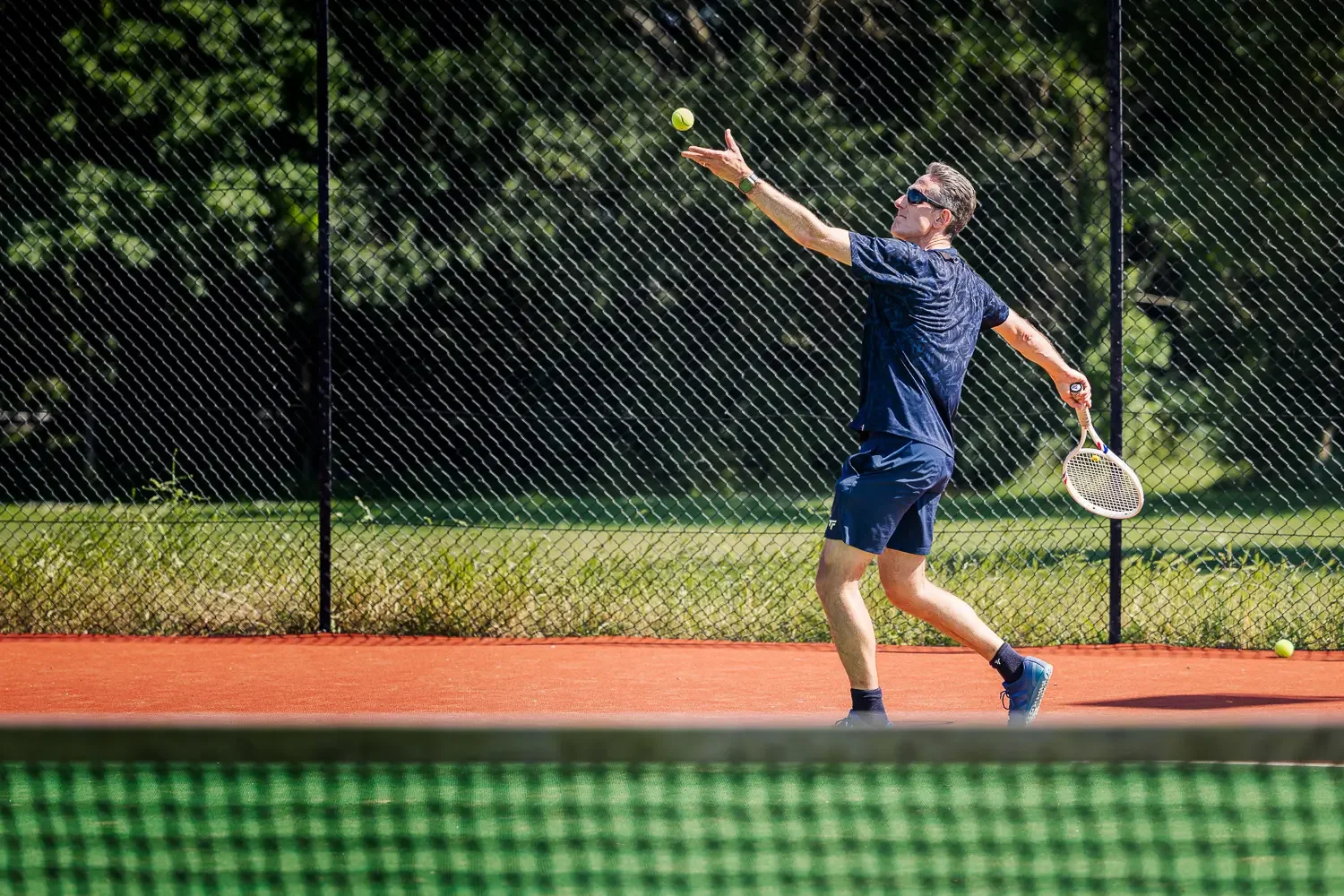 Tennis coach Howard mid-serve, tossing the ball on a clay court at Greenwood Park Lawn Tennis Club