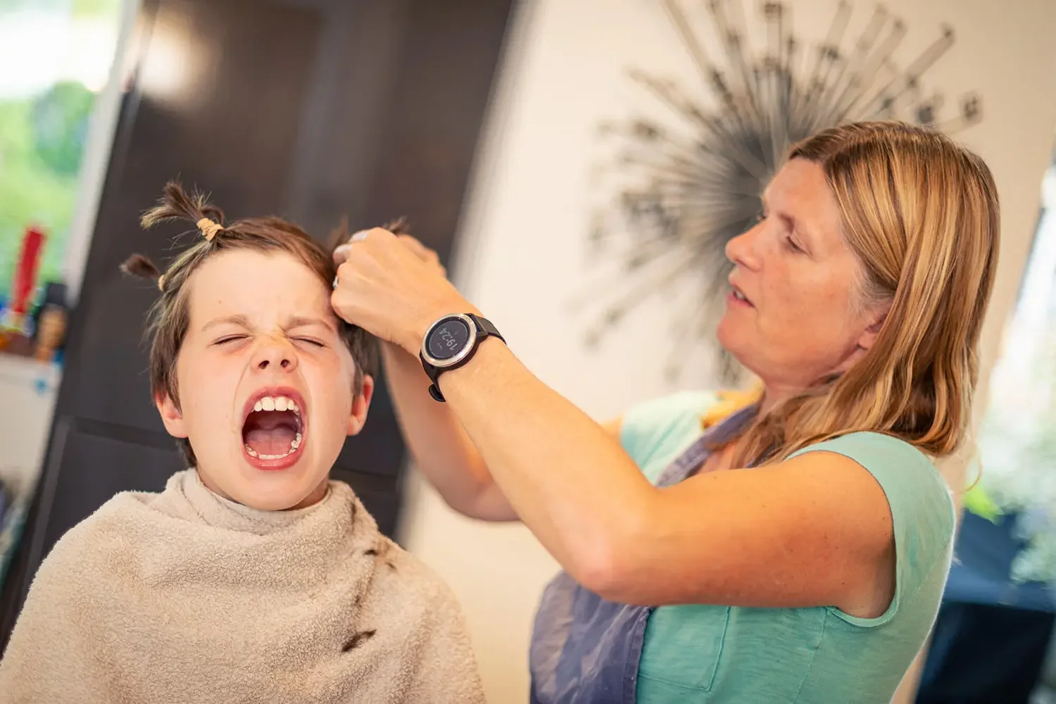 A woman shaving a young boy's hair in a home setting, with the boy making a grimace.