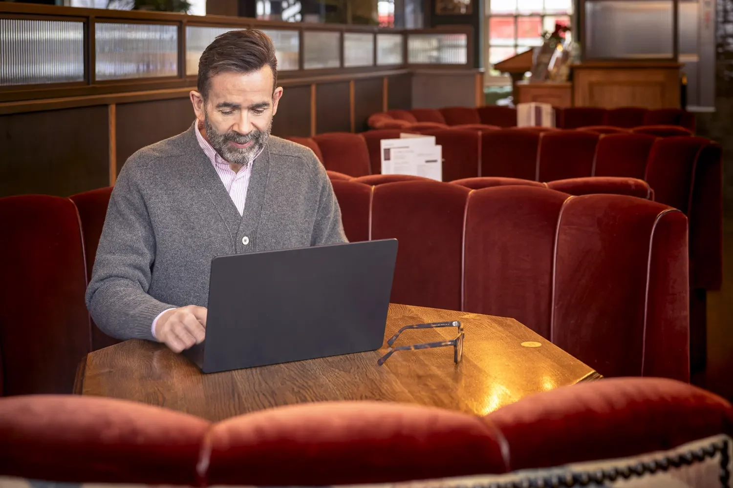 A middle-aged man with a beard and glasses, wearing a gray sweater over a pink collared shirt, sitting alone at a wooden table with a laptop in a cozy restaurant with red upholstered booths and wood-paneled walls.