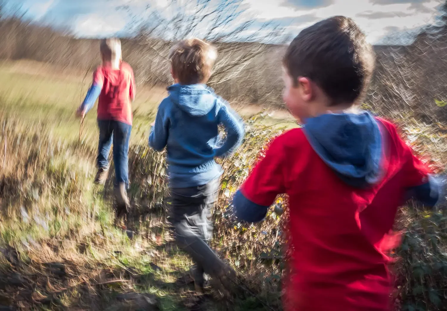 Three children running outdoors during daytime on a trail with grassy and shrub-covered terrain, with a cloudy sky.