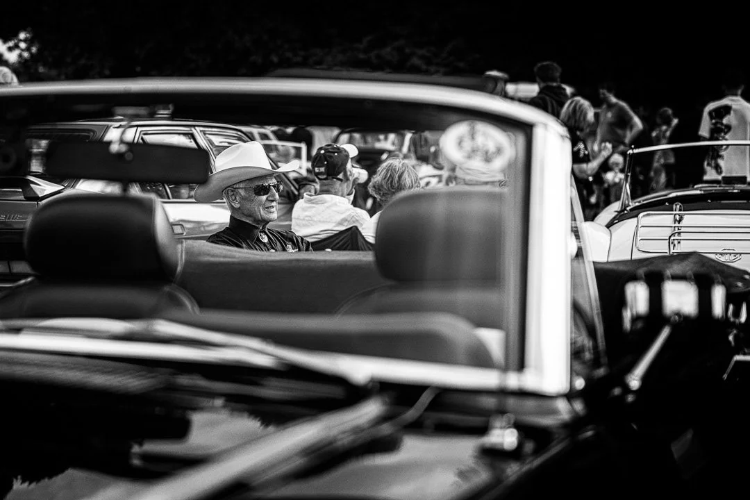 A man wearing a stetson hat and sunglasses sitting beside a classic car.jpg