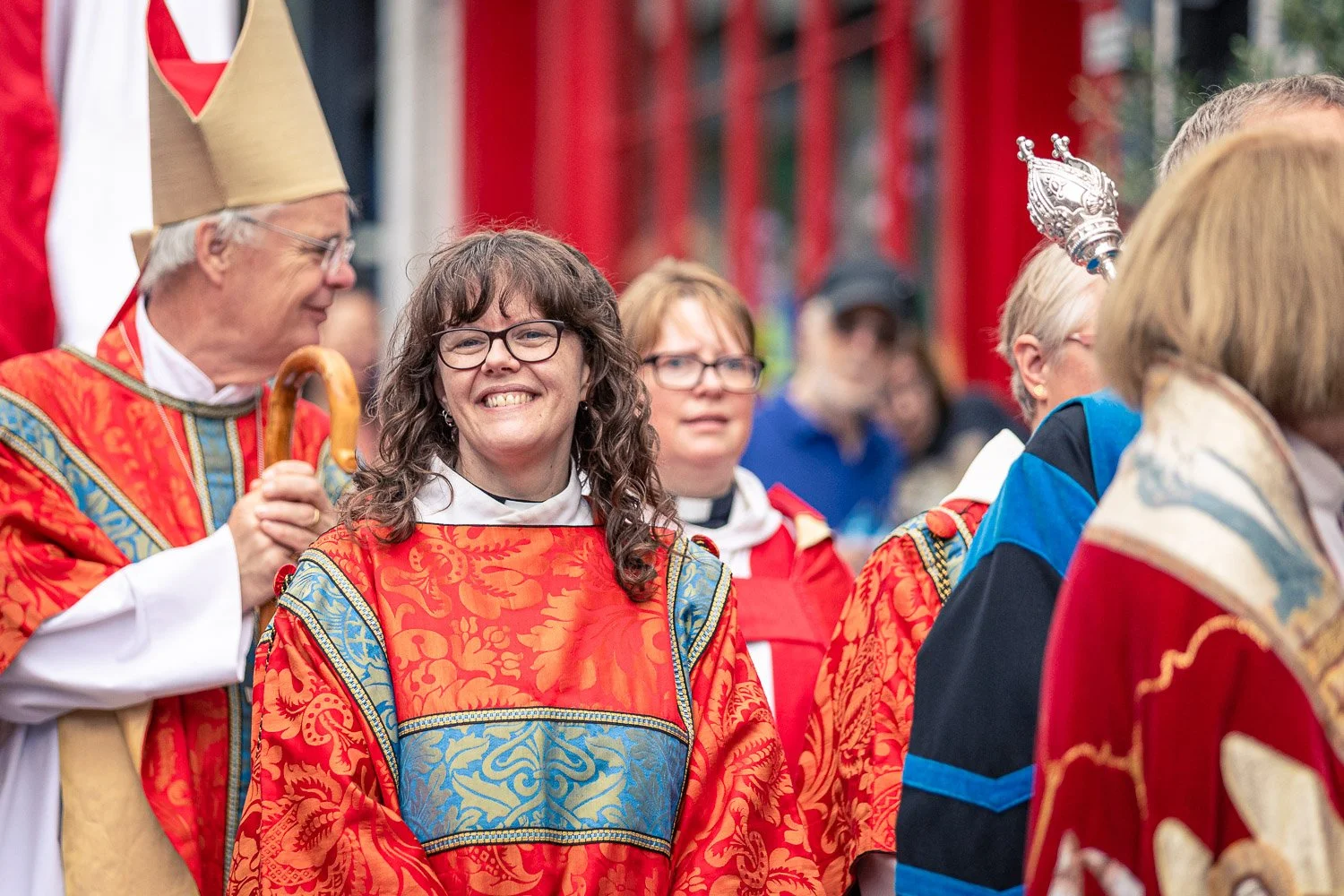  Smiling female clergy member in red and gold vestments during the Alban Day 2026 pilgrimage procession in St Albans, Hertfordshire, with the Bishop of St Albans visible behind her. 