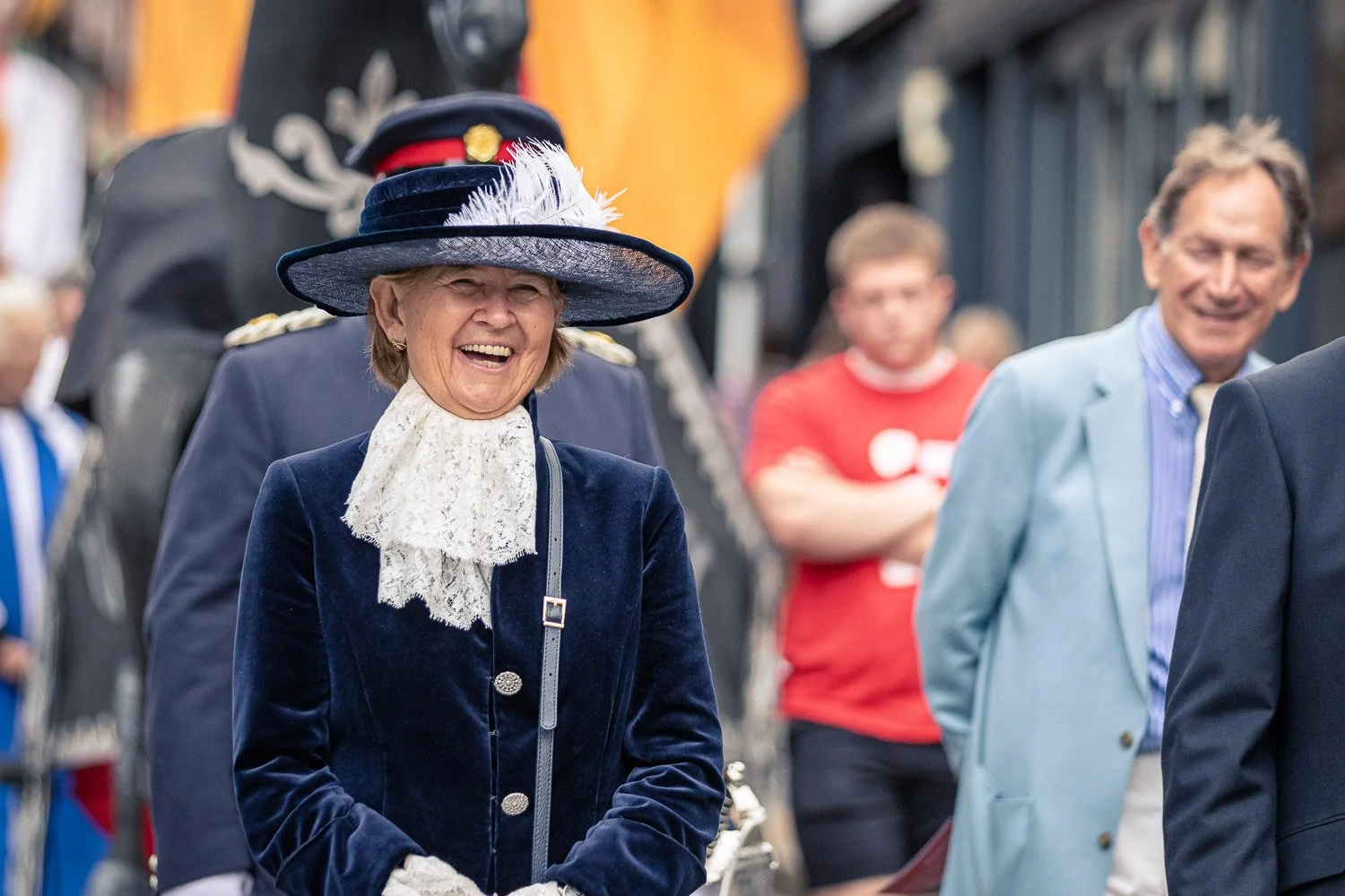  Woman in navy blue ceremonial dress and feathered hat laughing during the Alban Day 2026 procession through St Albans, Hertfordshire. 