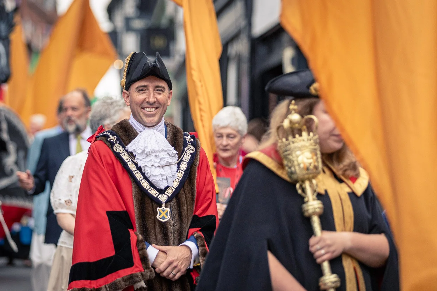  The Mayor of St Albans in red ceremonial robes and chain of office smiling during the Alban Day 2026 procession, with orange flags and a mace bearer visible, Hertfordshire. 