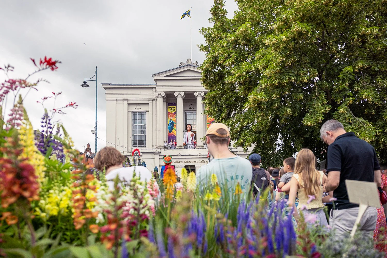  Crowd watching Alban Day 2026 celebrations from a flower-filled garden in front of St Albans Museum and Gallery, Hertfordshire, with a performer on the steps. 