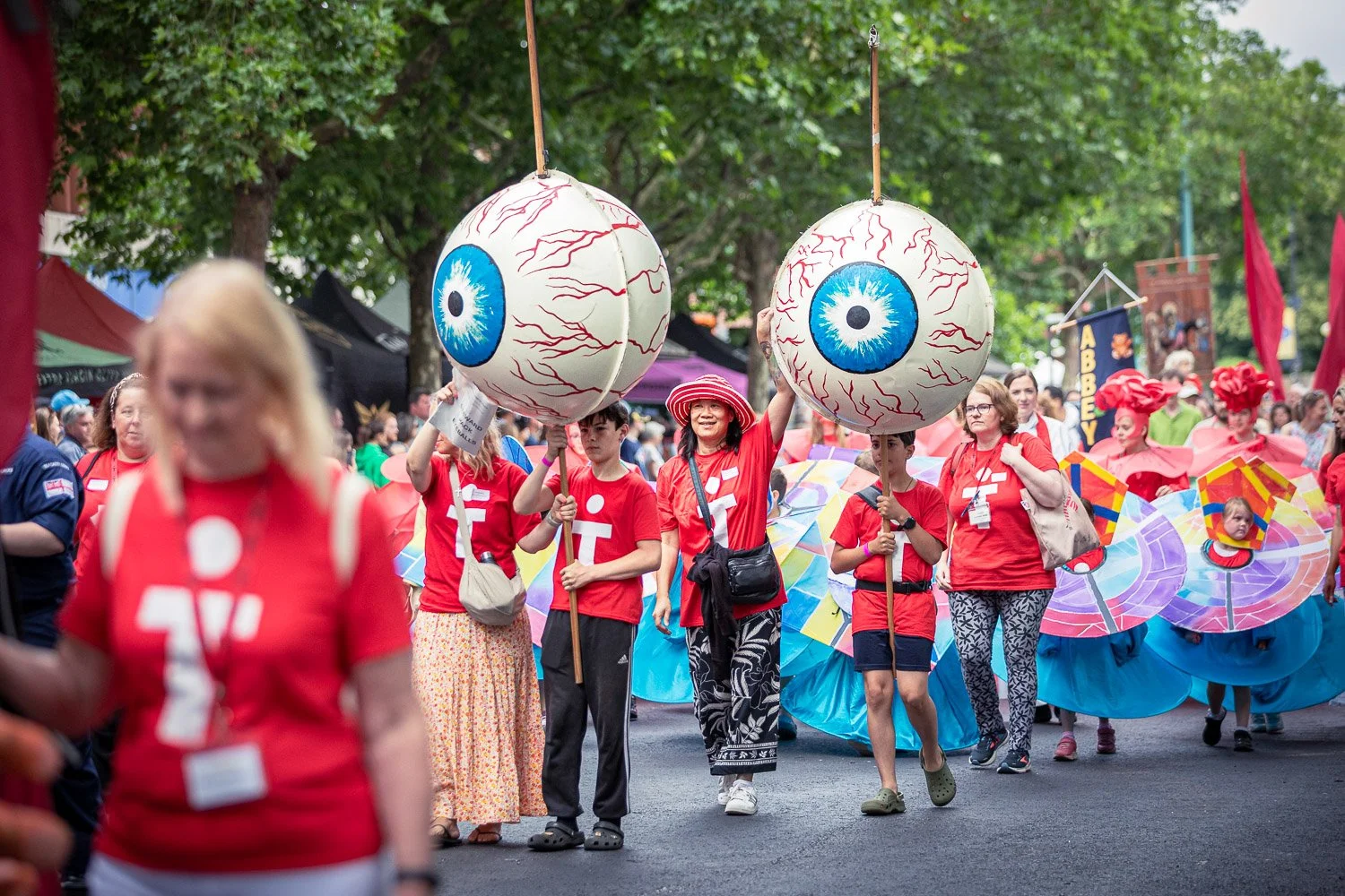  Pilgrims in red T-shirts carrying giant eyeball props on poles during the Alban Day 2026 procession in St Albans, Hertfordshire, with colourful carnival costumes behind them. 