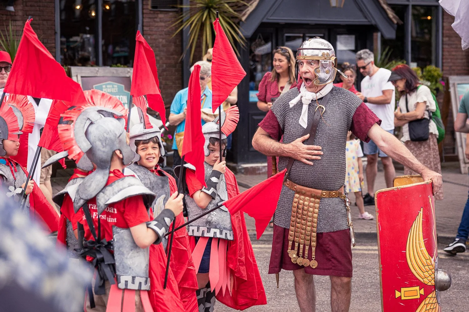  Roman soldier reenactor in full armour reacting with children dressed as Roman soldiers carrying red flags during the Alban Day 2026 procession in St Albans, Hertfordshire. 