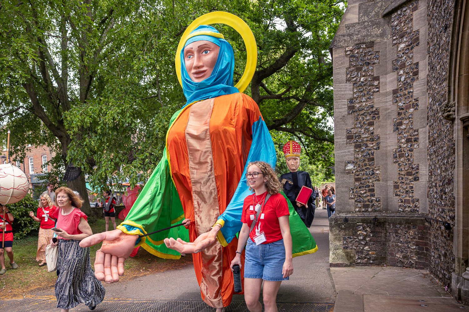  Large colourful saint puppet with blue veil and yellow halo processing through the grounds of St Albans Cathedral during Alban Day 2026, Hertfordshire, with a second bishop puppet visible behind. 