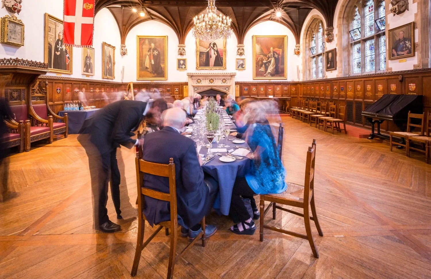 Guests at a long dinner table in a historic panelled hall, with waiting staff blurred in motion