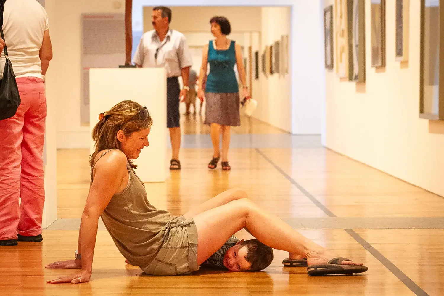 A woman sitting on the wooden floor of an art gallery with a young boy lying across her lap. The woman is smiling and looking down at the boy, who has his head resting on her leg, with other visitors walking in the background.