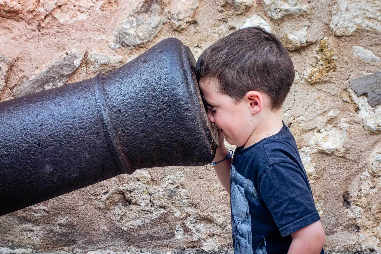 A young boy whispering into a large, old, black cannon against a textured stone wall background.
