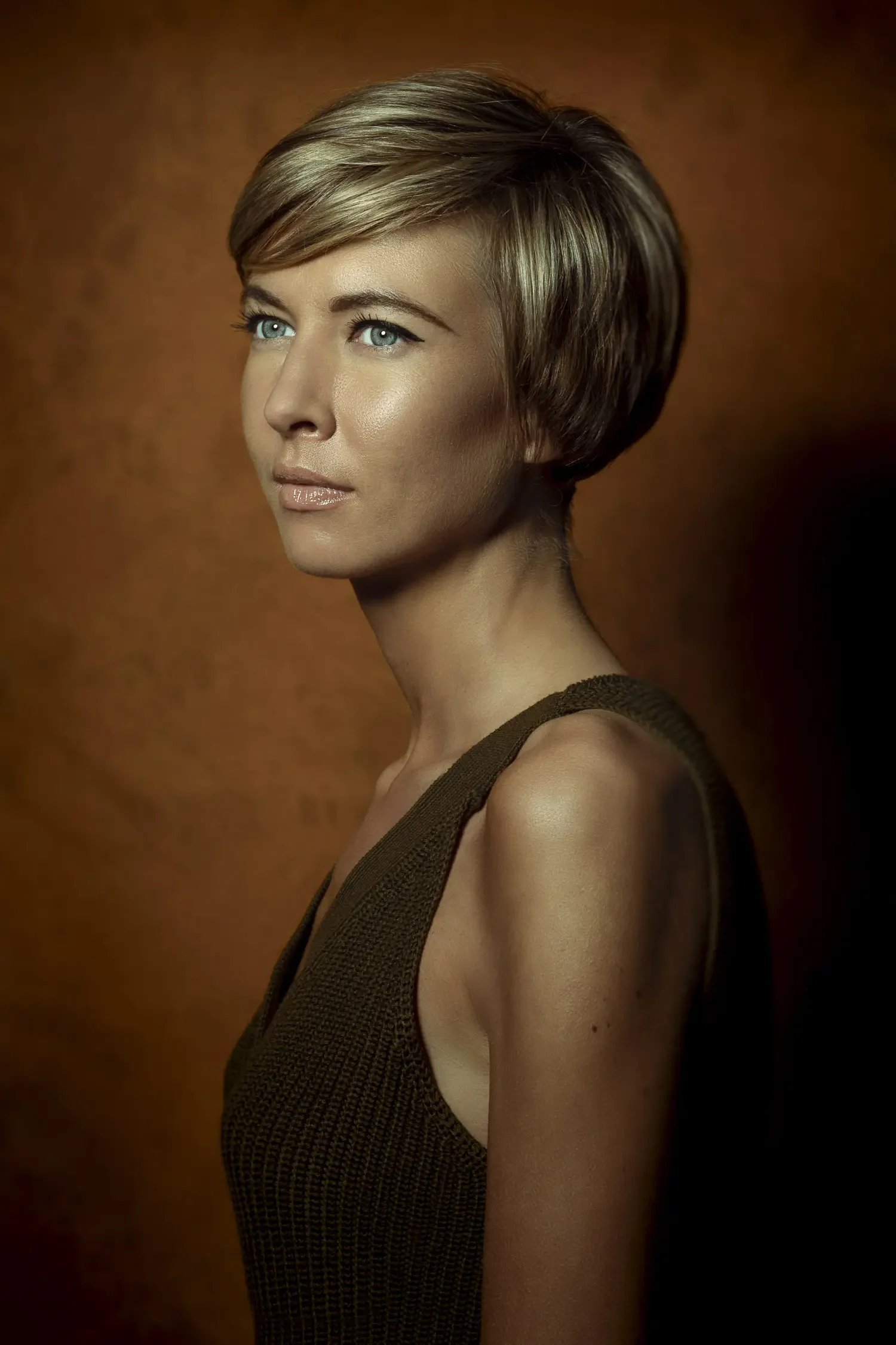 A woman with short blonde hair, blue eyes, light skin, and wearing a sleeveless brown top looking slightly to the left against a brown gradient background.