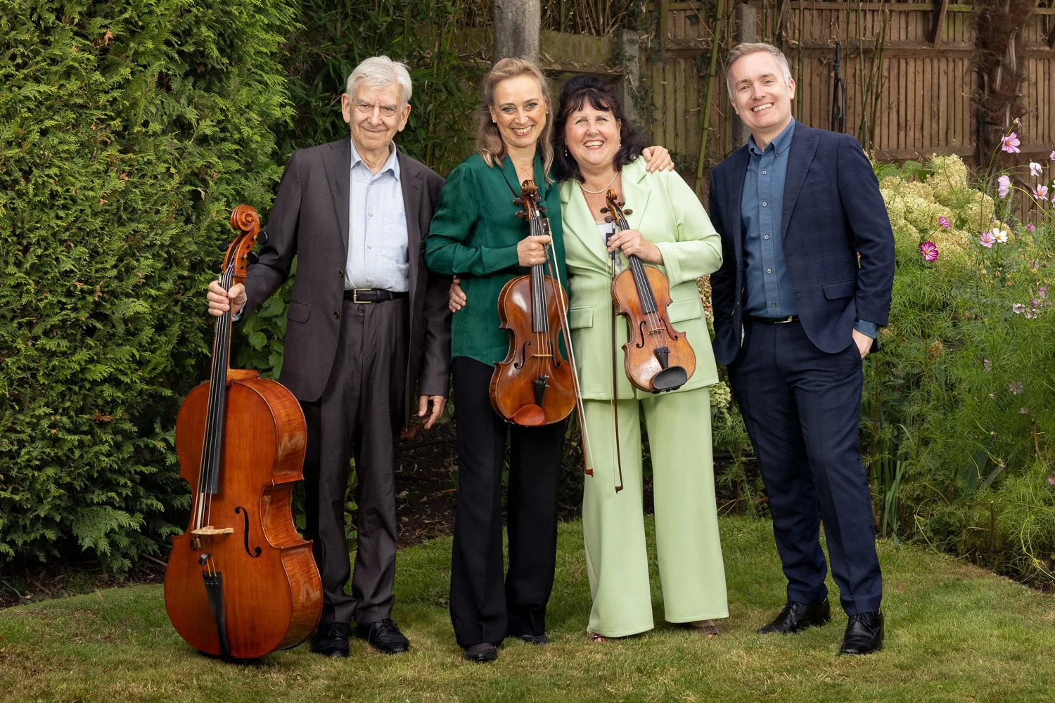 Four smiling classical musicians from The Zoffany Ensemble holding a cello, viola and violin in a garden in Arkley, near Barnet.
