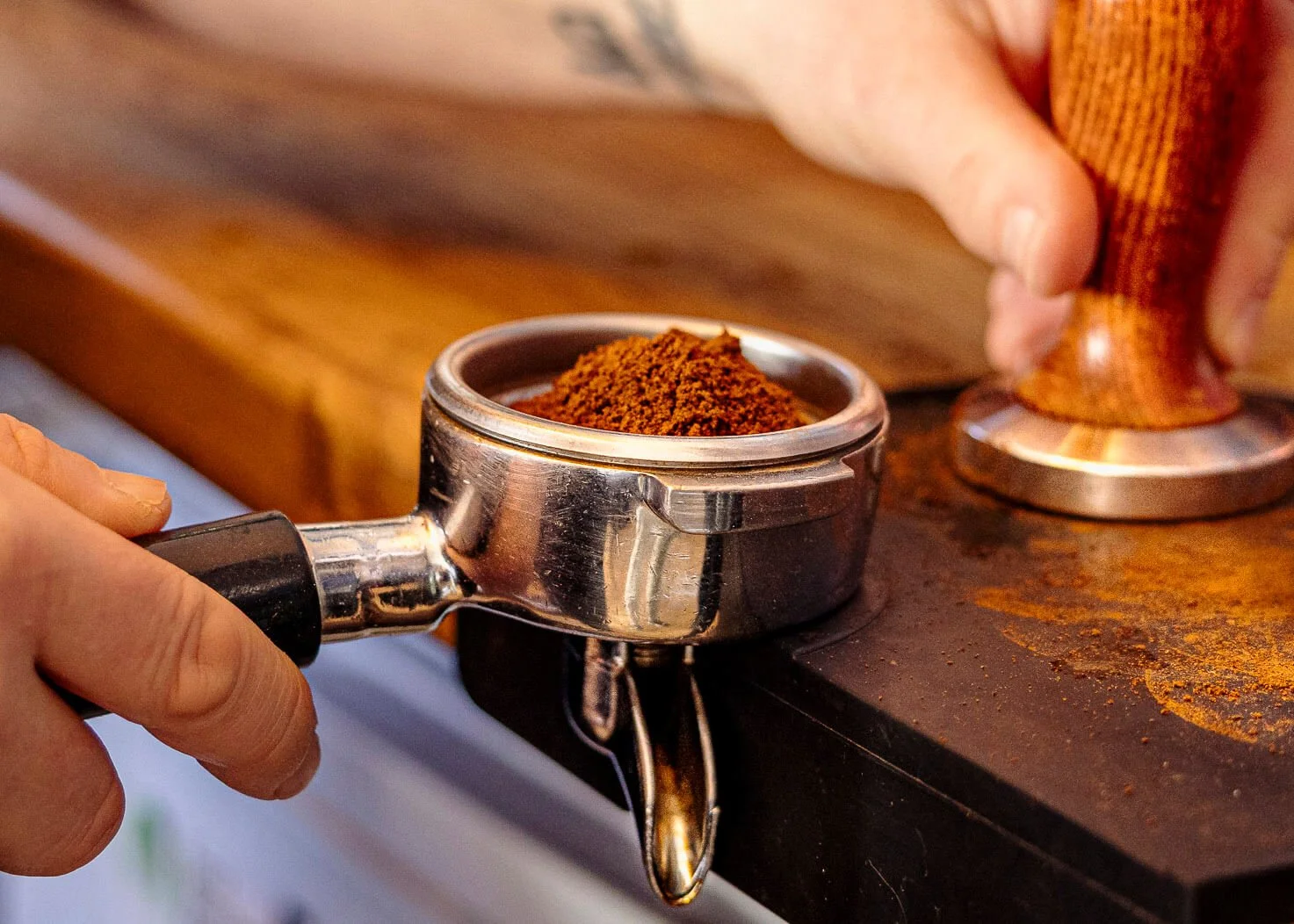 A coffee scoop filled with ground coffee on a wooden counter, with a person pressing down ground coffee with a tamper.